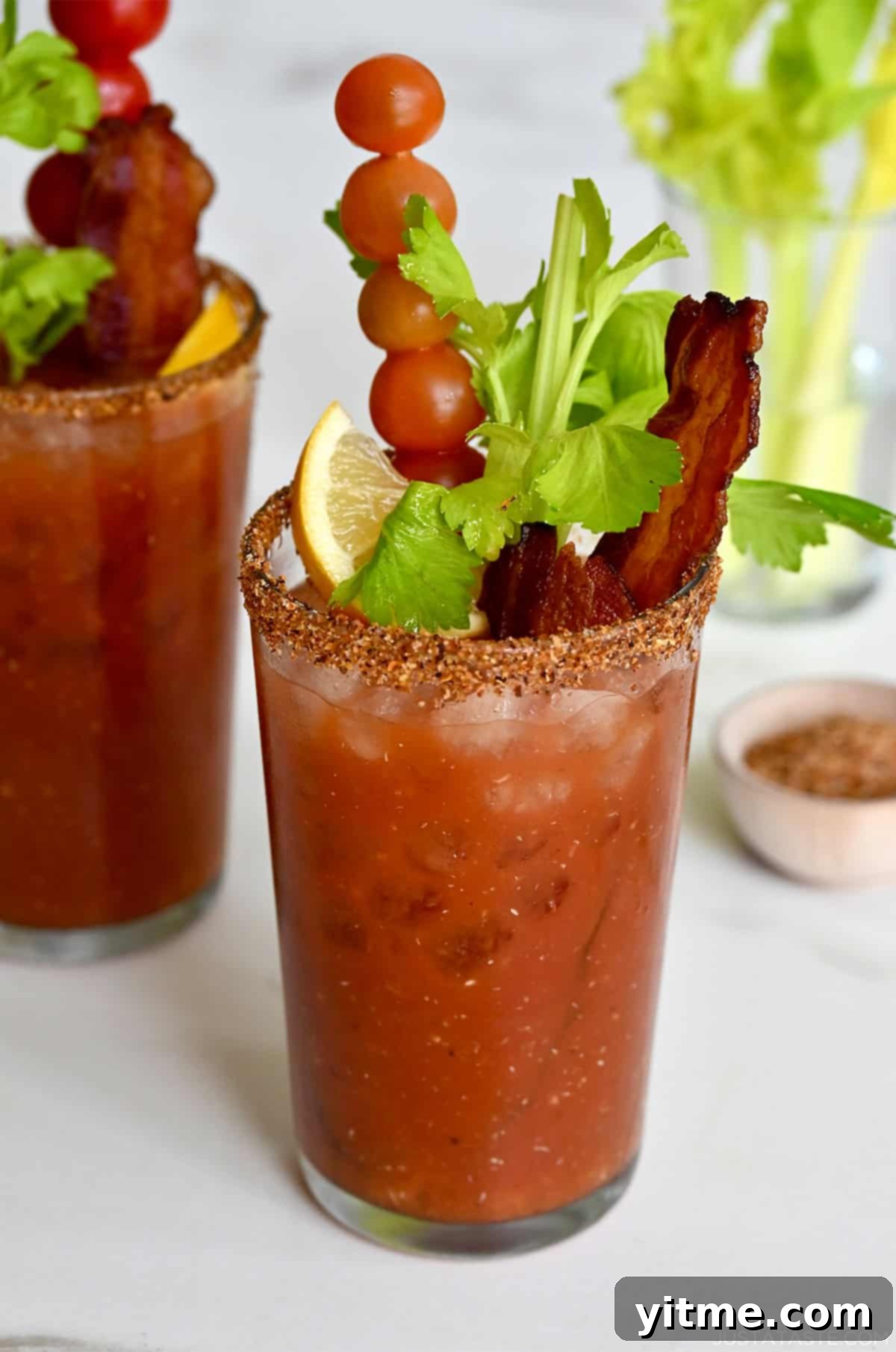 Bloody Mary Pitchers 8 Two tall glasses filled with homemade Bloody Mary mix and garnished with crispy bacon, cherry tomatoes on a skewer, celery sticks and a lemon wedge.