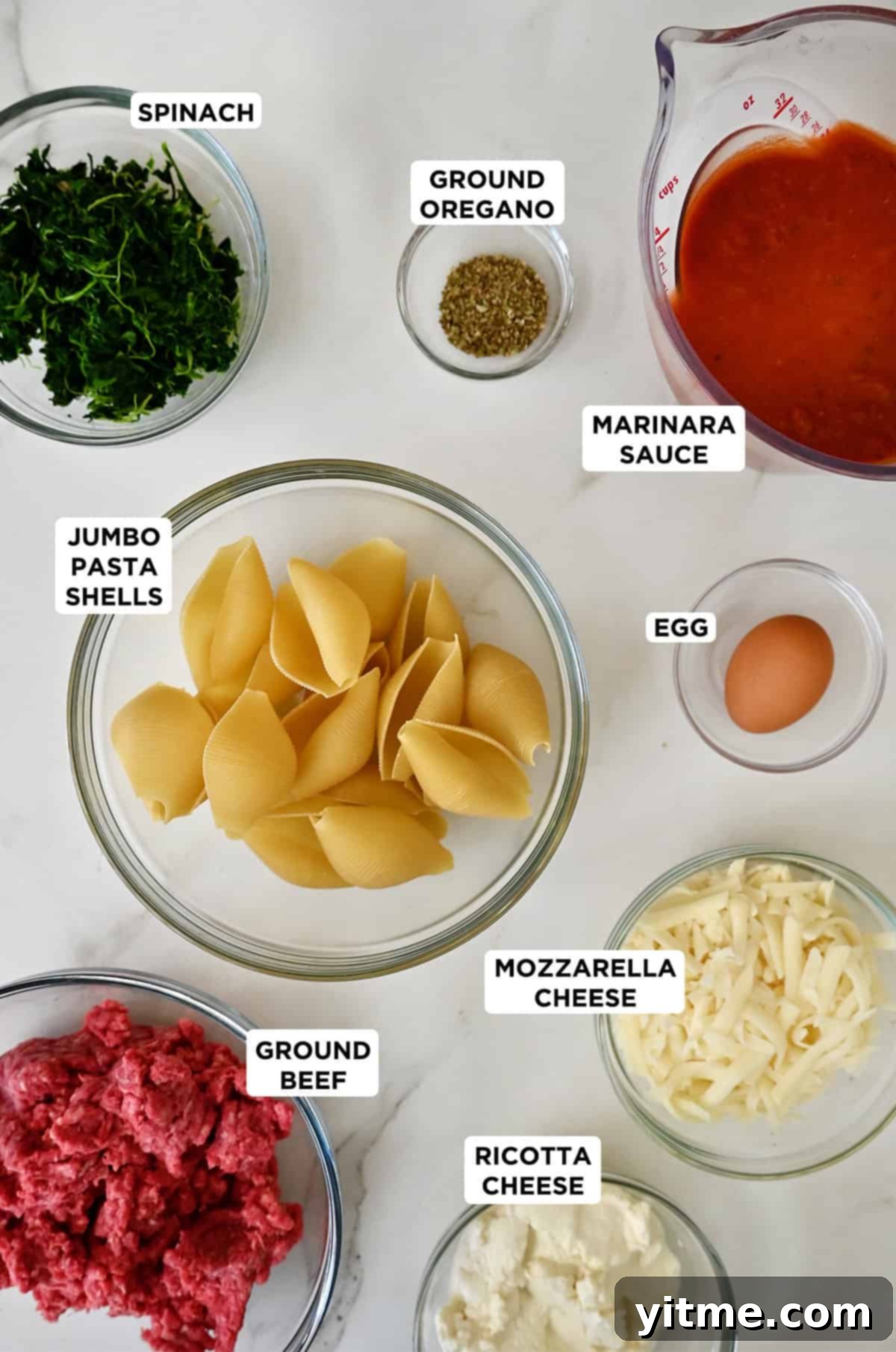 Stuffed shells ingredients in various sizes of glass bowls: jumbo pasta shells, spinach, dried oregano, marinara sauce, egg, mozzarella cheese, ricotta cheese and ground beef.