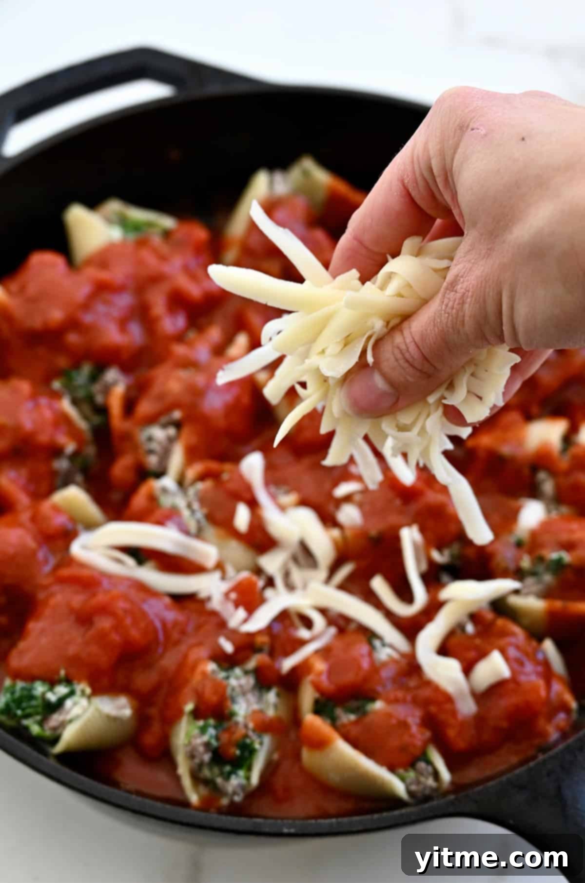 Adding shredded cheese and marinara sauce atop stuffed pasta shells in a baking dish.