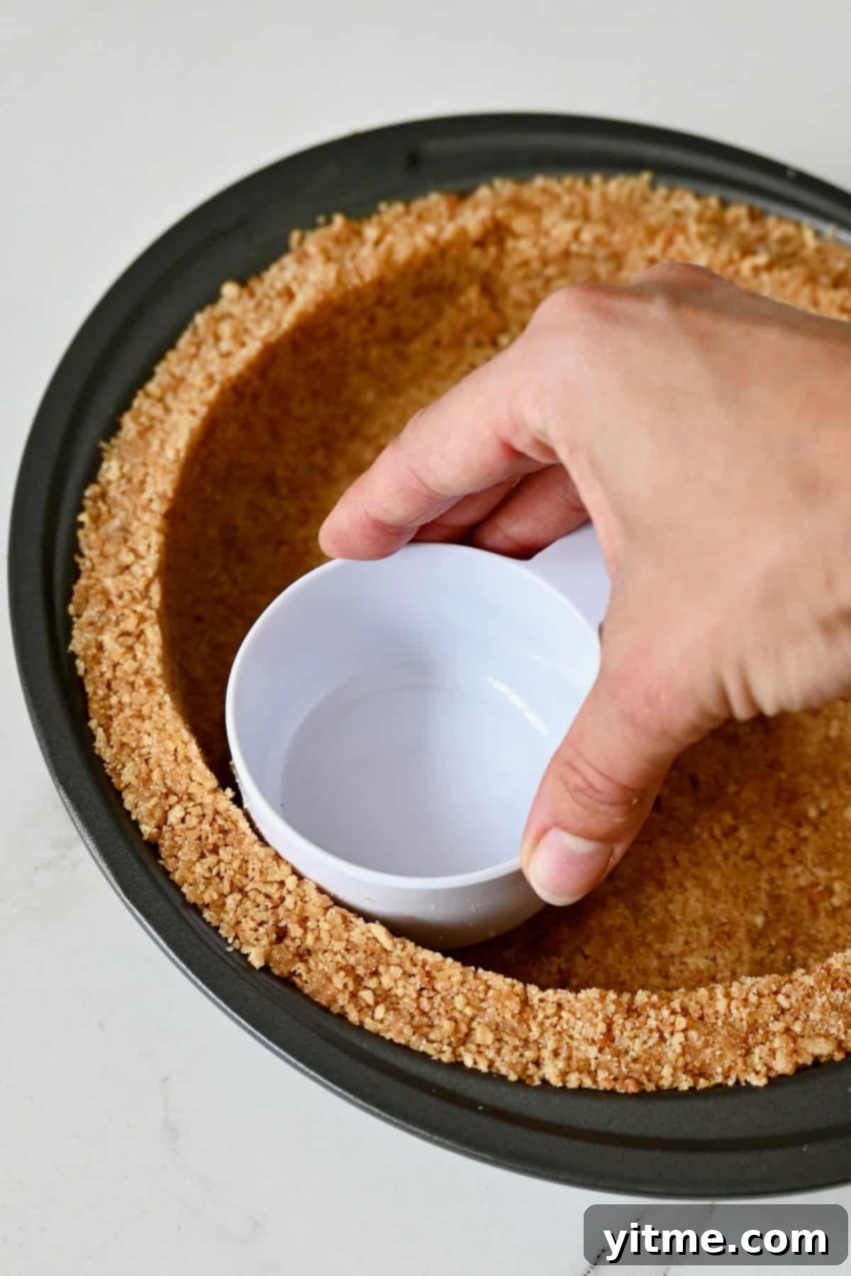 Mystery Ingredient Ice Cream Pie 8 A hand uses a measuring cup to firmly press waffle cone crumbs into a pie plate to form a compact, even crust.