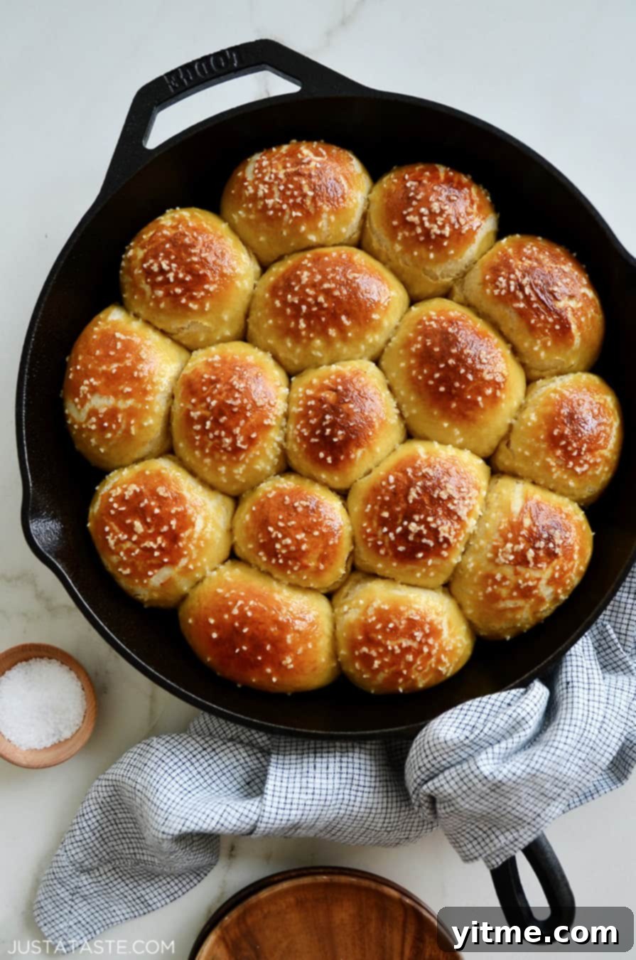 A rustic cast-iron skillet generously filled with freshly baked, golden-brown pull-apart pretzel rolls, with a small ceramic bowl of coarse salt on the side.
