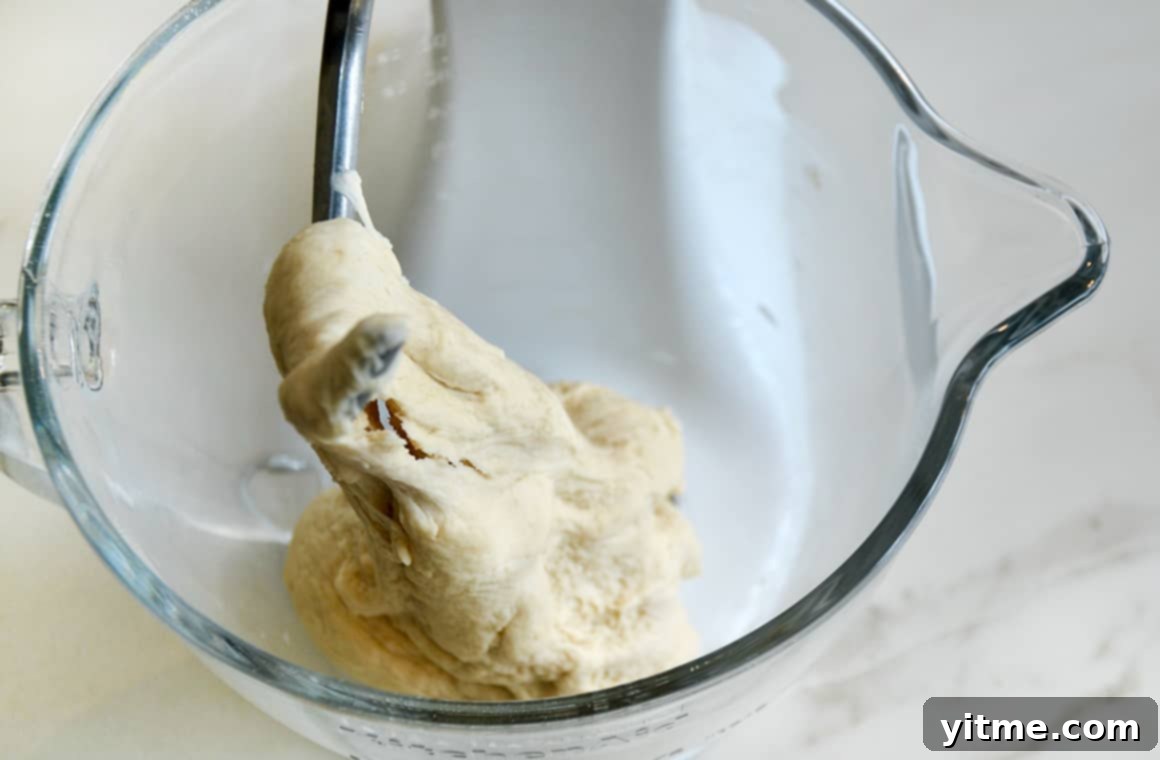 Freshly mixed pretzel dough wrapped around a dough hook in a clear glass mixing bowl, ready for its first rise.
