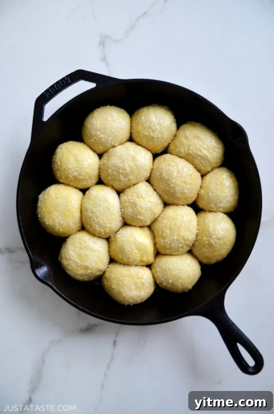 Unbaked pretzel roll dough balls, brushed with egg wash and topped with coarse salt, nestled snugly in a greased cast-iron skillet, ready for the oven.