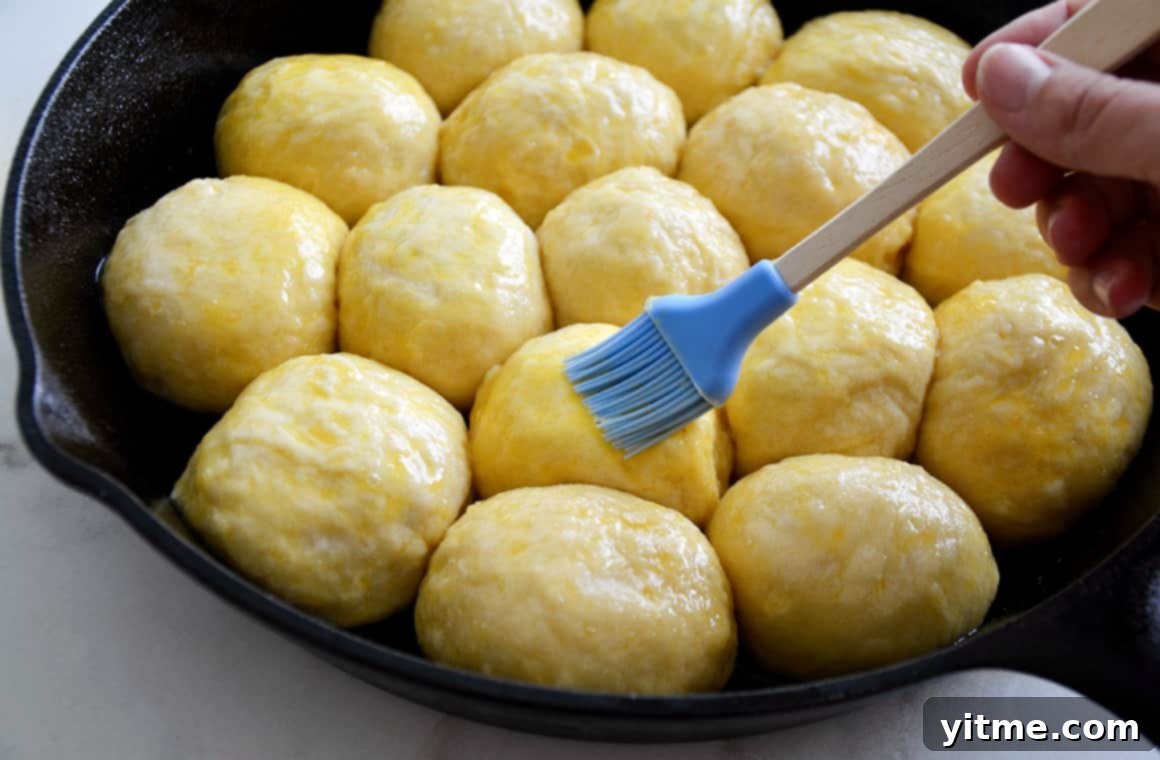 Pretzel dough balls arranged in a cast-iron skillet are being brushed with an egg wash, a crucial step for a shiny, golden-brown crust.