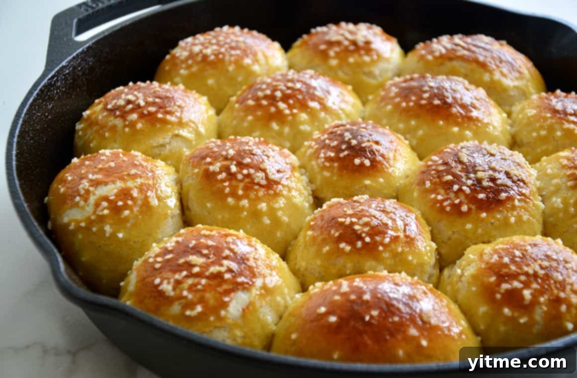 A freshly baked cast-iron skillet filled with warm, golden-brown soft pretzel rolls, generously topped with coarse salt.