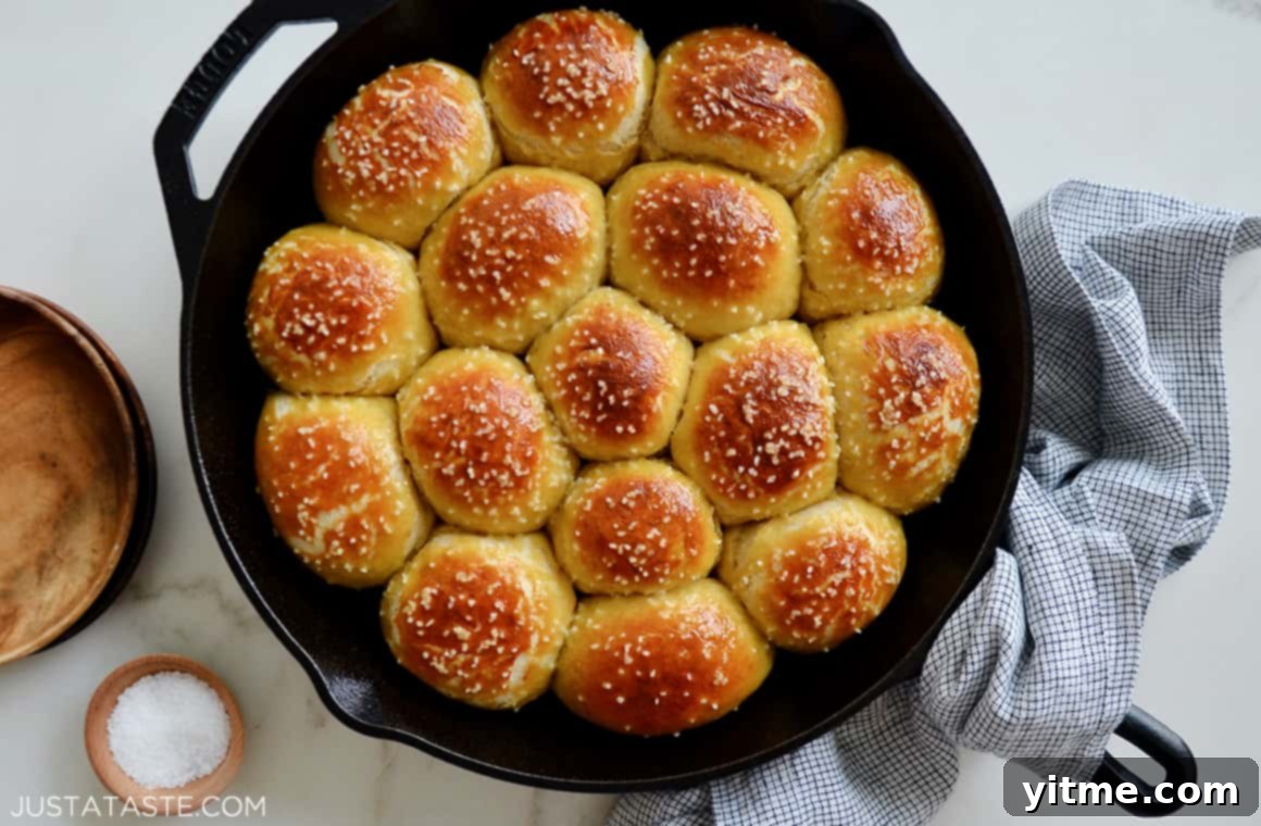 A cast-iron skillet brimming with freshly baked, golden-brown pull-apart pretzel rolls, with a beige napkin elegantly wrapped around the handle.