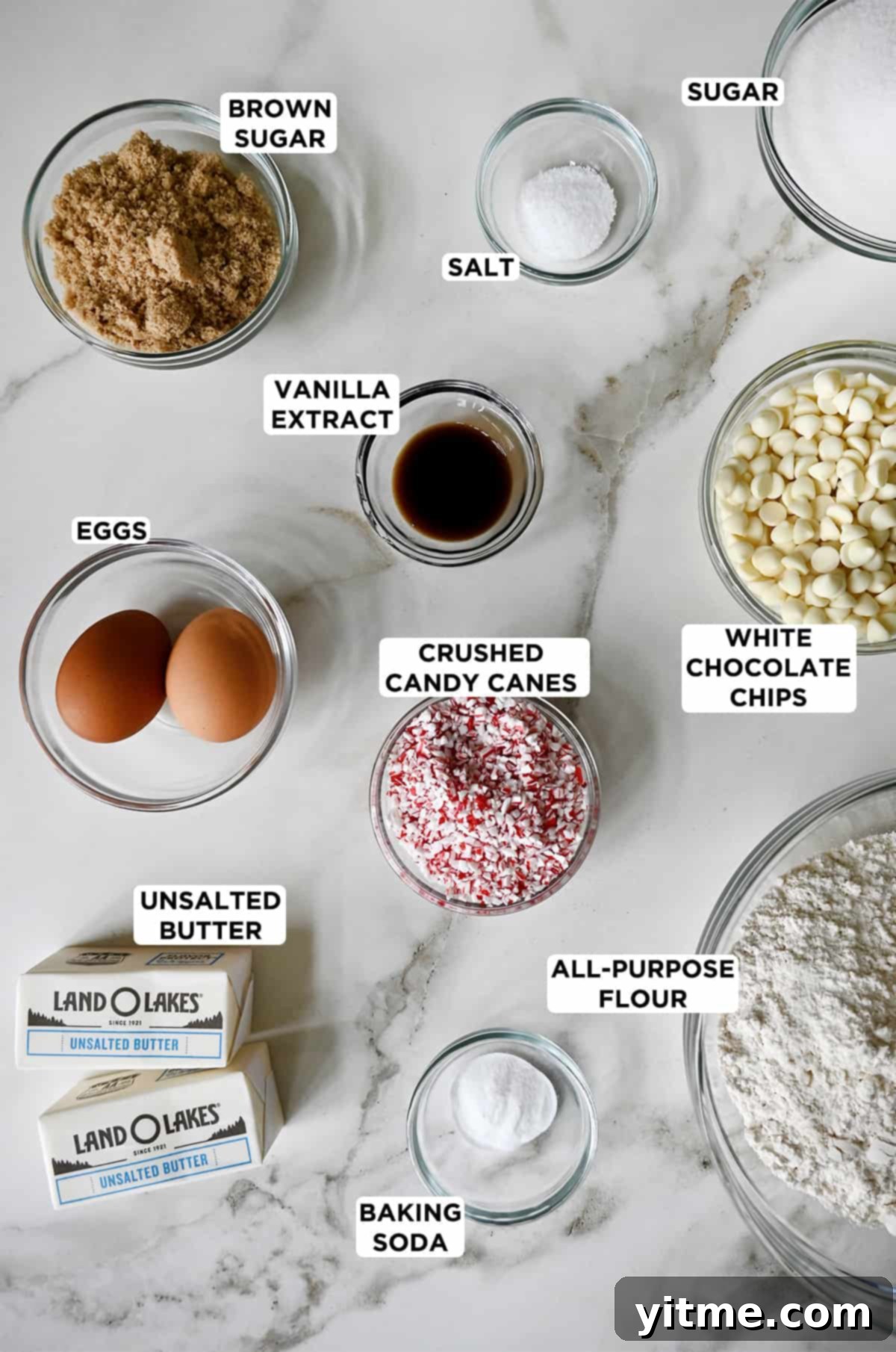 A colorful array of ingredients for peppermint white chocolate cookies in clear bowls, including light brown sugar, salt, white sugar, vanilla extract, white chocolate chips, crushed candy canes, all-purpose flour, baking soda, butter, and eggs, ready for baking.