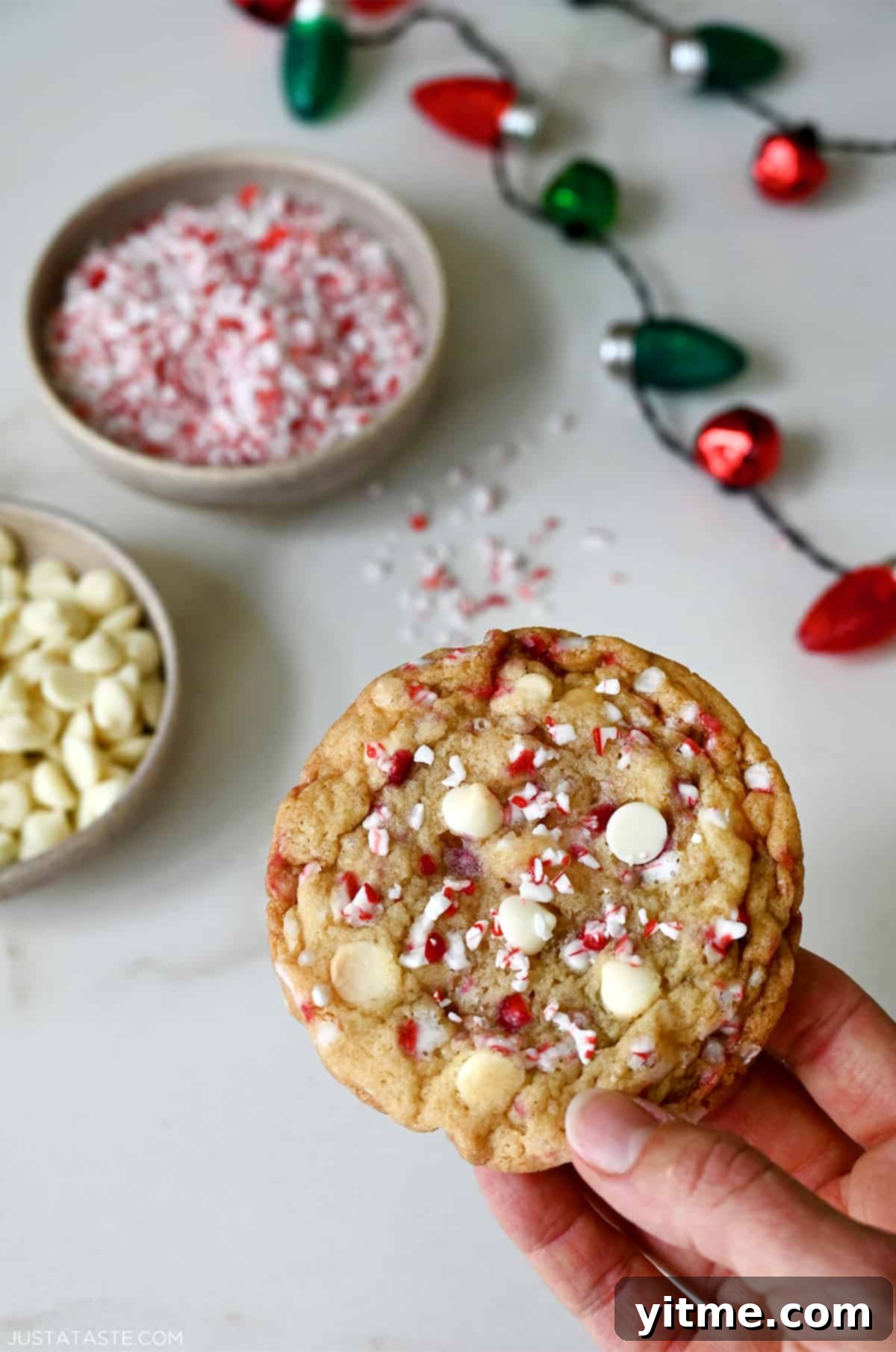 A hand holds a freshly baked white chocolate peppermint cookie, beautifully studded with white chocolate chips and vibrant crushed candy canes, showcasing its soft, chewy texture.