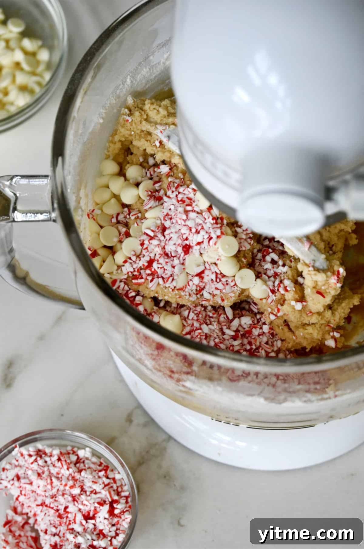 A clear bowl in a white stand mixer holds cookie dough, generously topped with an abundant mix of crushed candy canes and white chocolate chips, with an additional small bowl of crushed candy canes beside it.