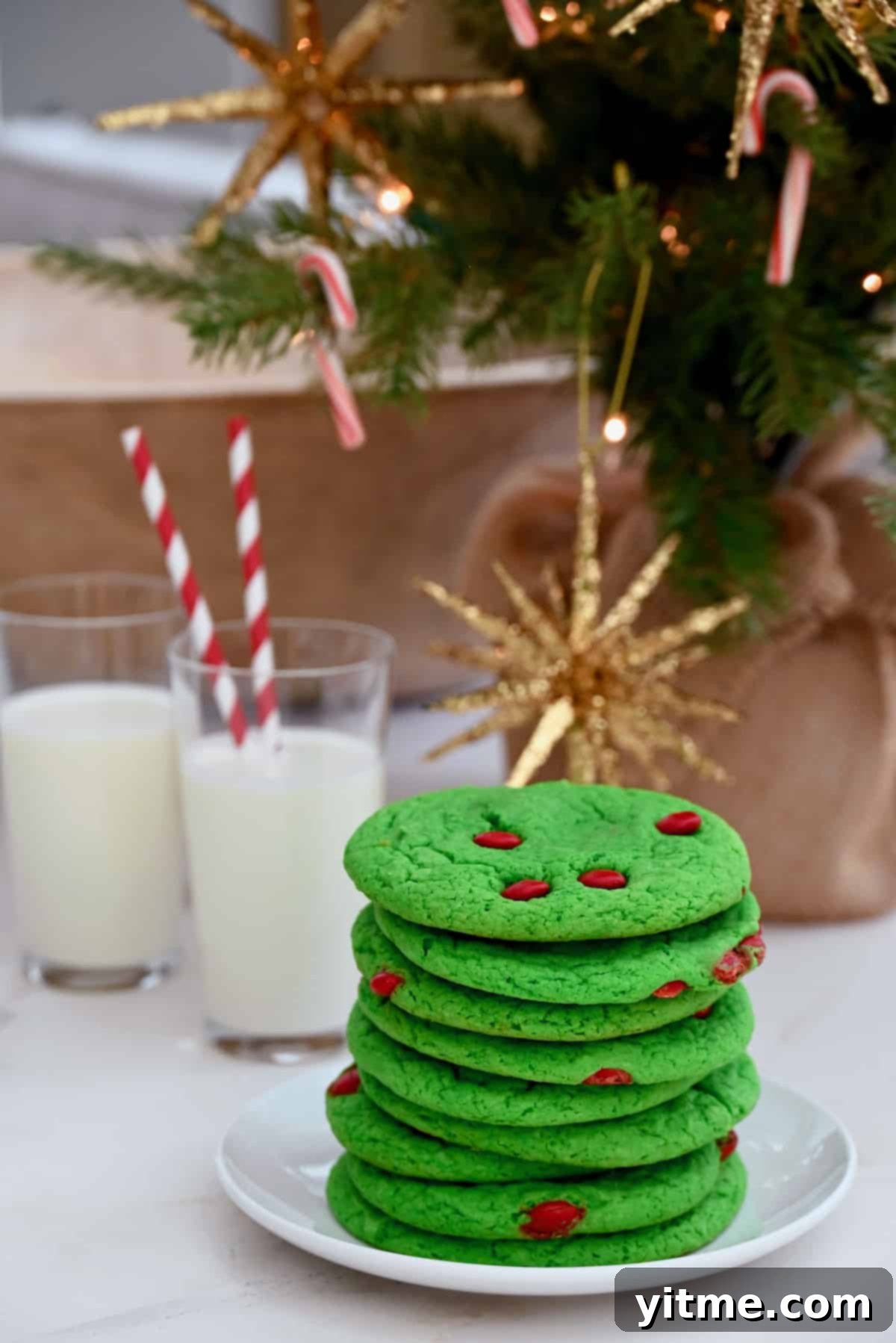 Green cake mix cookies with red M&Ms are stacked in front of glasses of milk with red striped straws. A Christmas tree is in the background.