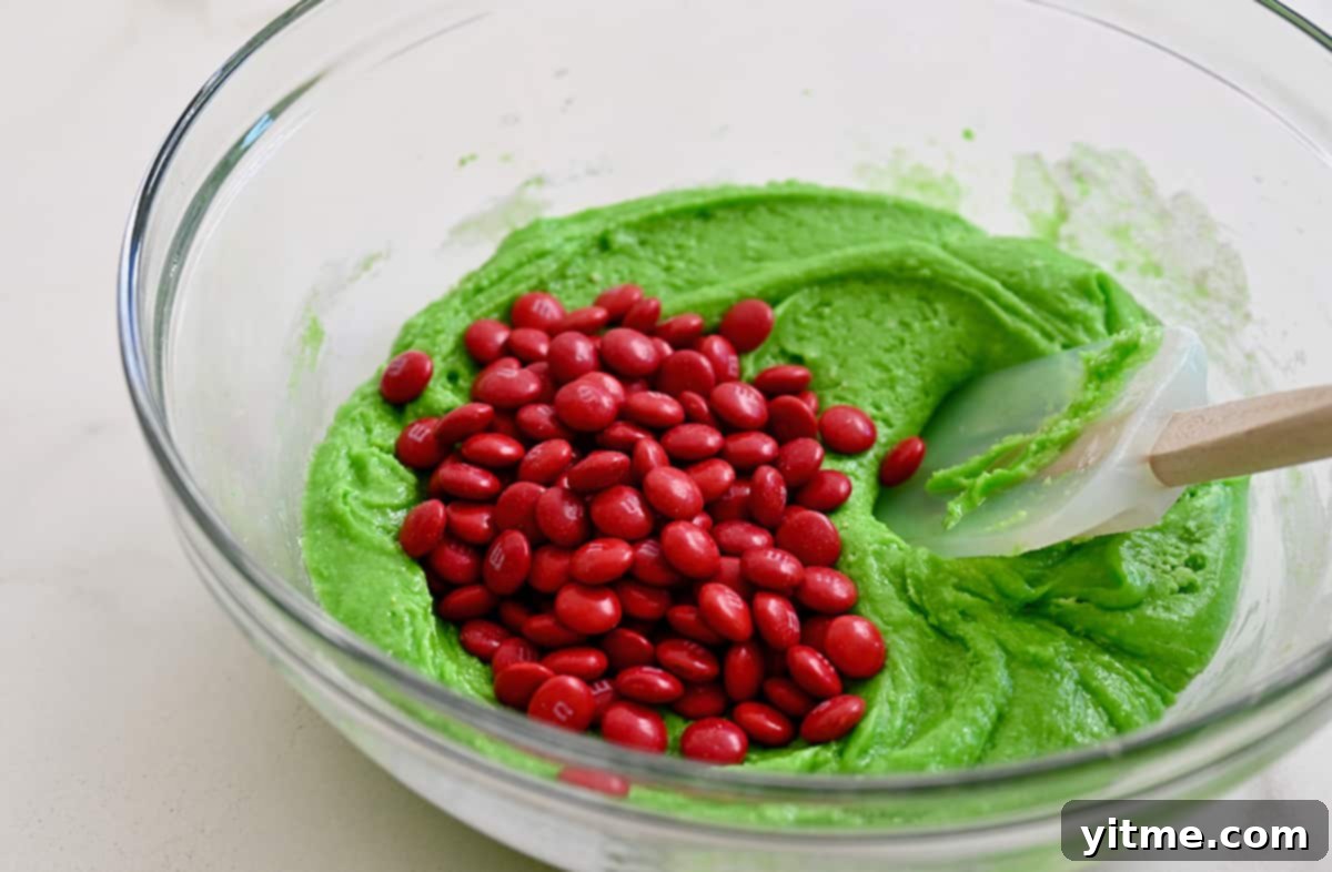 A glass bowl holds bright-green cake mix batter with red M&Ms ready to be stirred in with a spatula.
