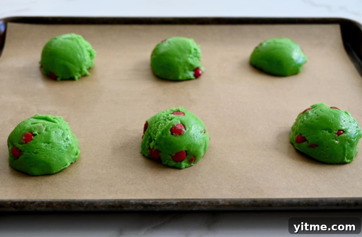 Balls of green cake mix cookie dough with red M&Ms are spaced out on a baking sheet lined with parchment paper.