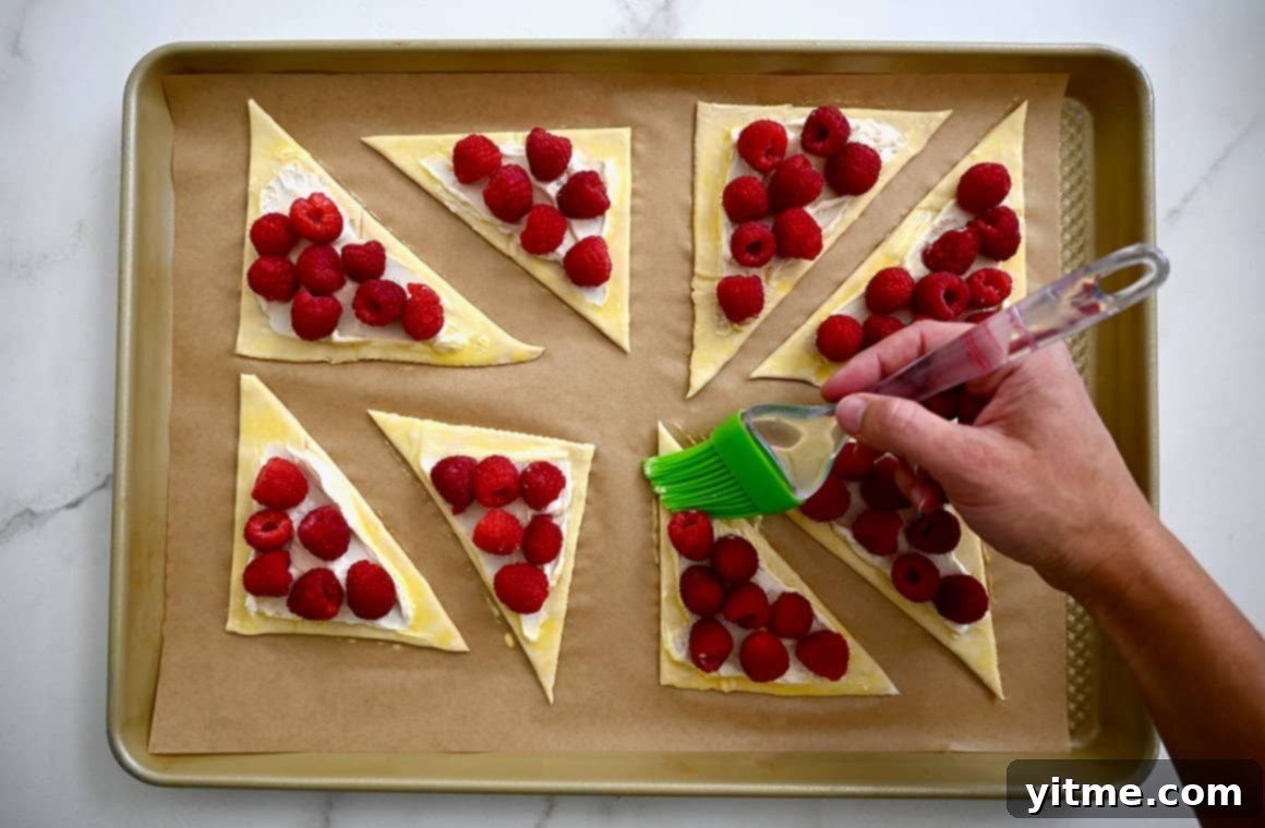 Raspberry Cream Cheese Danish 5 A hand holding a pastry brush applies an egg wash to triangle-shaped danish on a parchment paper-lined baking sheet