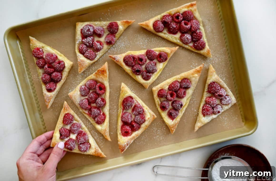 Raspberry Cream Cheese Danish 6 A hand reaches for a cream cheese danish with raspberries that's dusted with confectioners' sugar