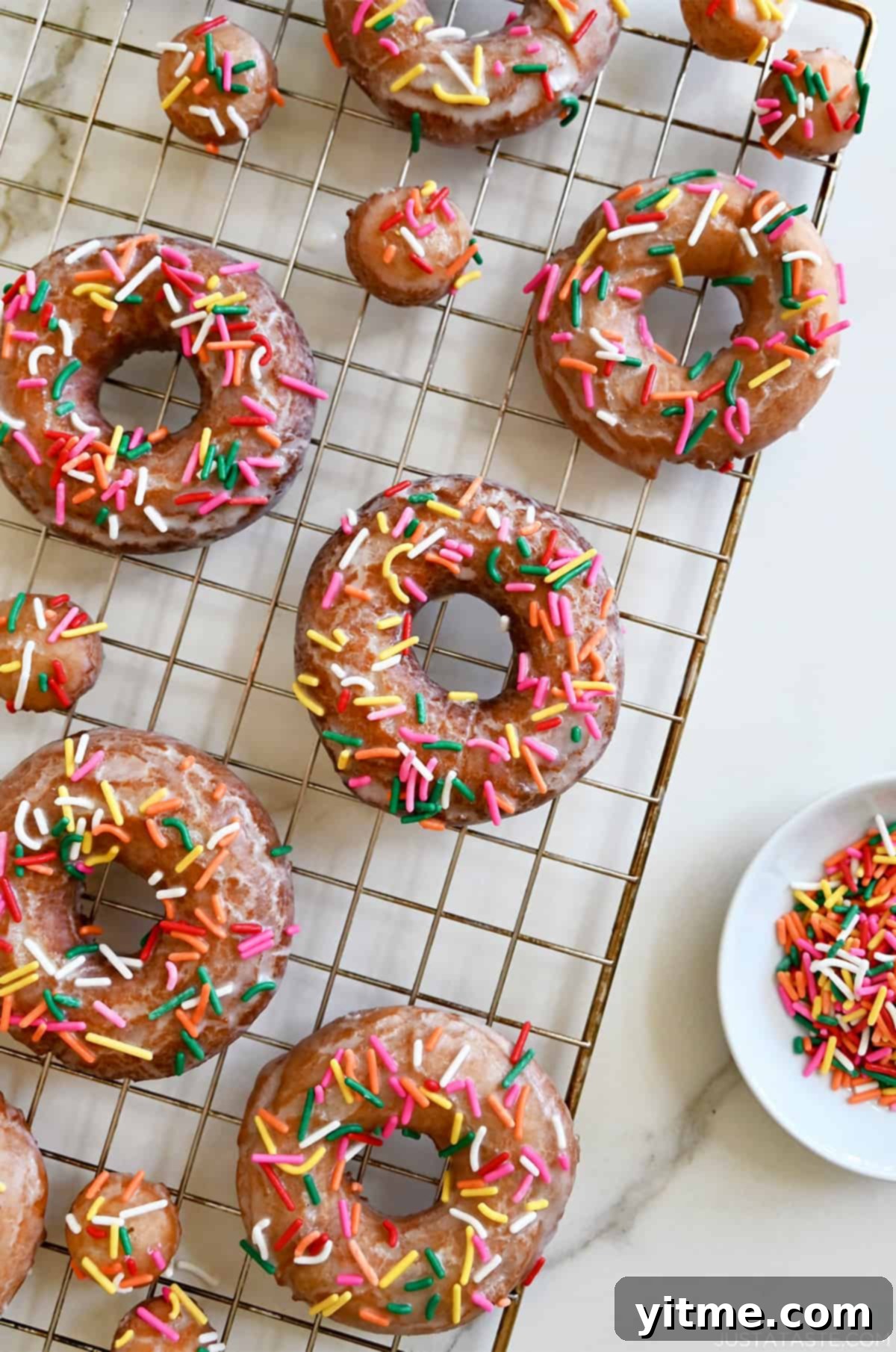 Sour cream doughnuts coated in a vanilla glaze and topped with rainbow sprinkles on a wire cooling rack.