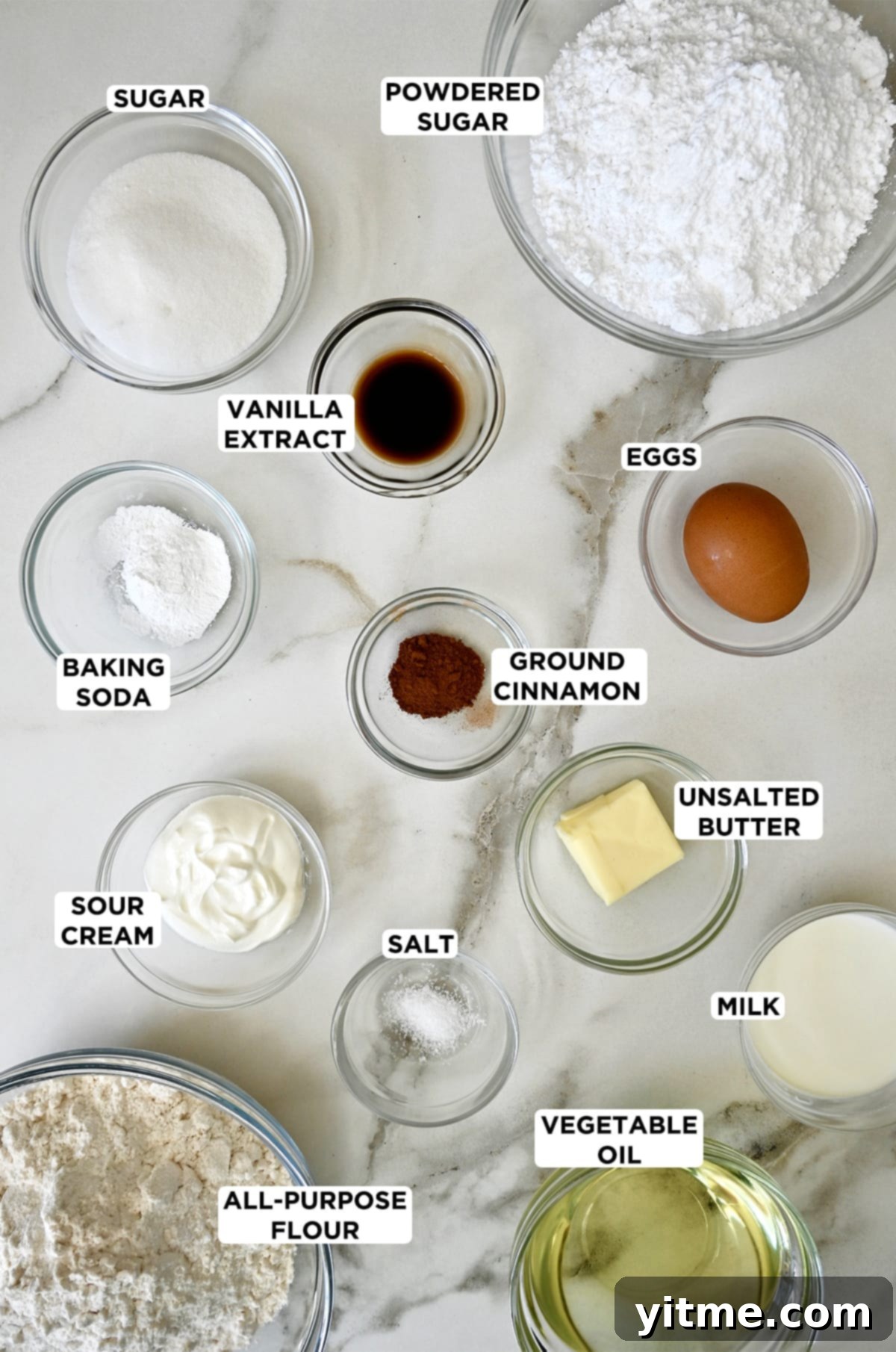 Various sizes of clear bowl containing powdered sugar, an egg, unsalted butter, milk, vegetable oil, salt, flour, sour cream, baking soda, vanilla extract and granulated sugar.