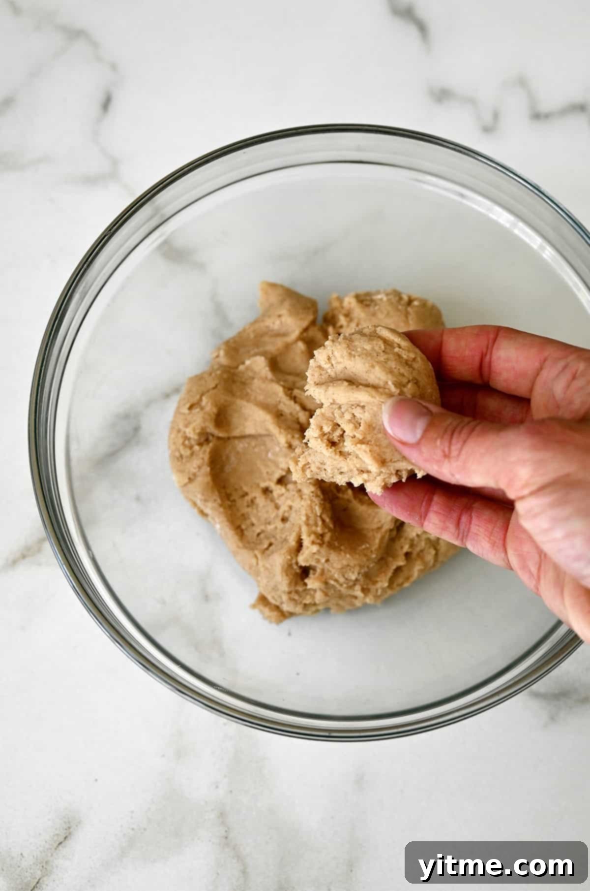 A hand holds a clump of doughnut dough over a bowl containing dough.
