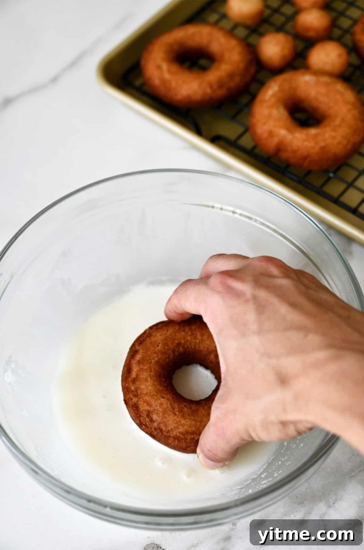 A hand holding a doughnut dips it into bowl with a homemade vanilla glaze. 