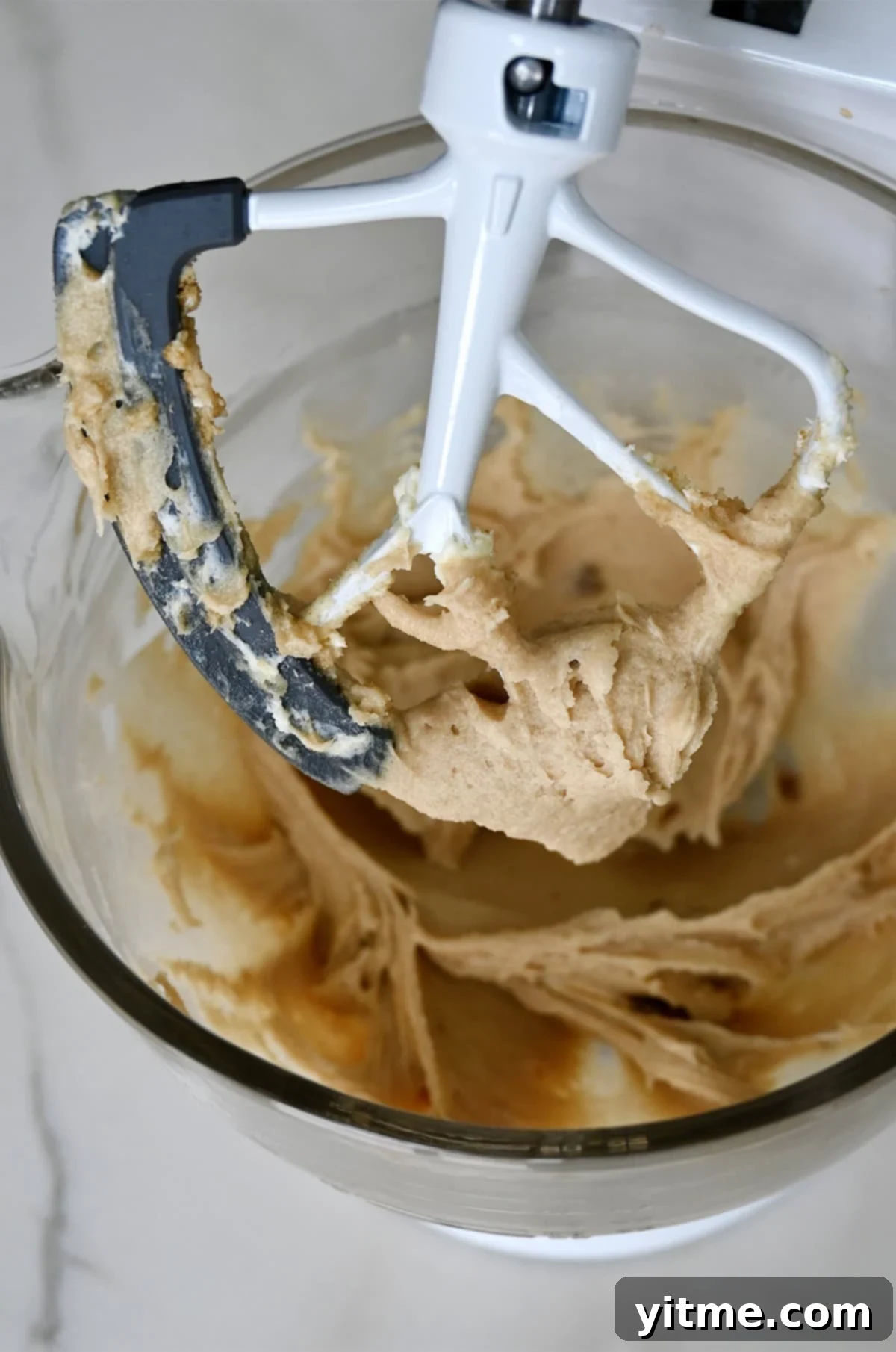 Creamed butter, cream cheese, white sugar, light brown sugar, and vanilla extract covering the paddle attachment of a stand mixer, indicating the first stage of cookie dough preparation.
