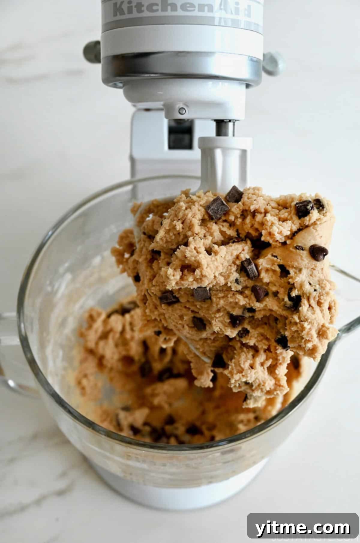 Rich chocolate chip cookie dough perfectly coats the paddle attachment of a stand mixer, indicating it's ready for scooping and baking.