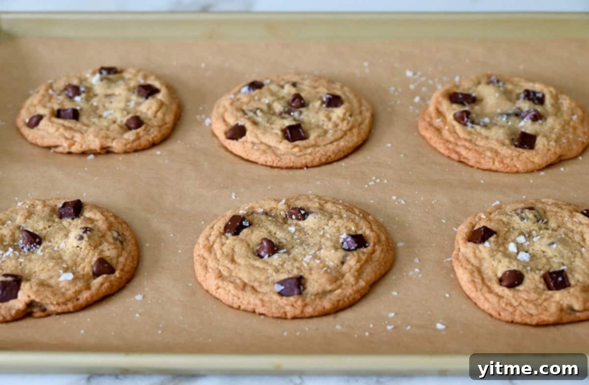 A close-up of warm, freshly baked soft chocolate chip cookies, still on the baking sheet, sprinkled with large-flake sea salt for enhanced flavor.