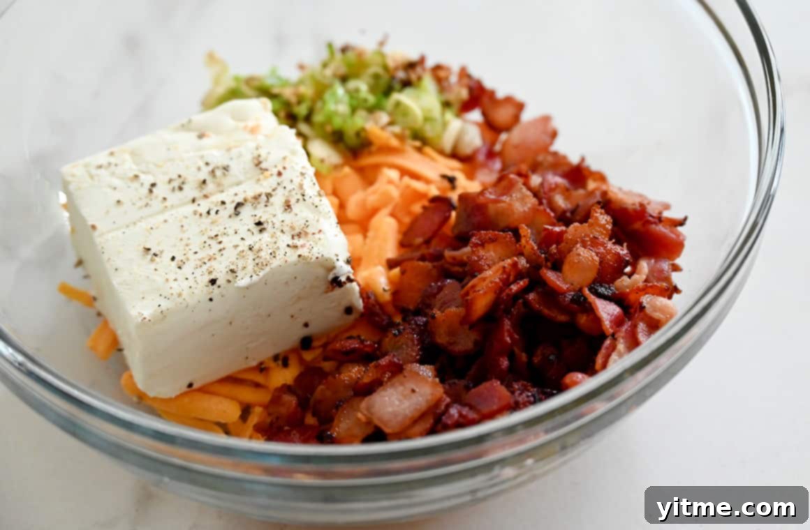 A clear bowl containing cream cheese, sliced scallions, shredded cheddar cheese and bacon pieces, ready for mixing