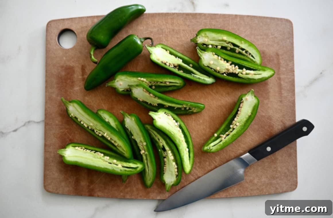 Halved jalapeños on a cutting board next to a sharp knife, with seeds and pith removed from some peppers