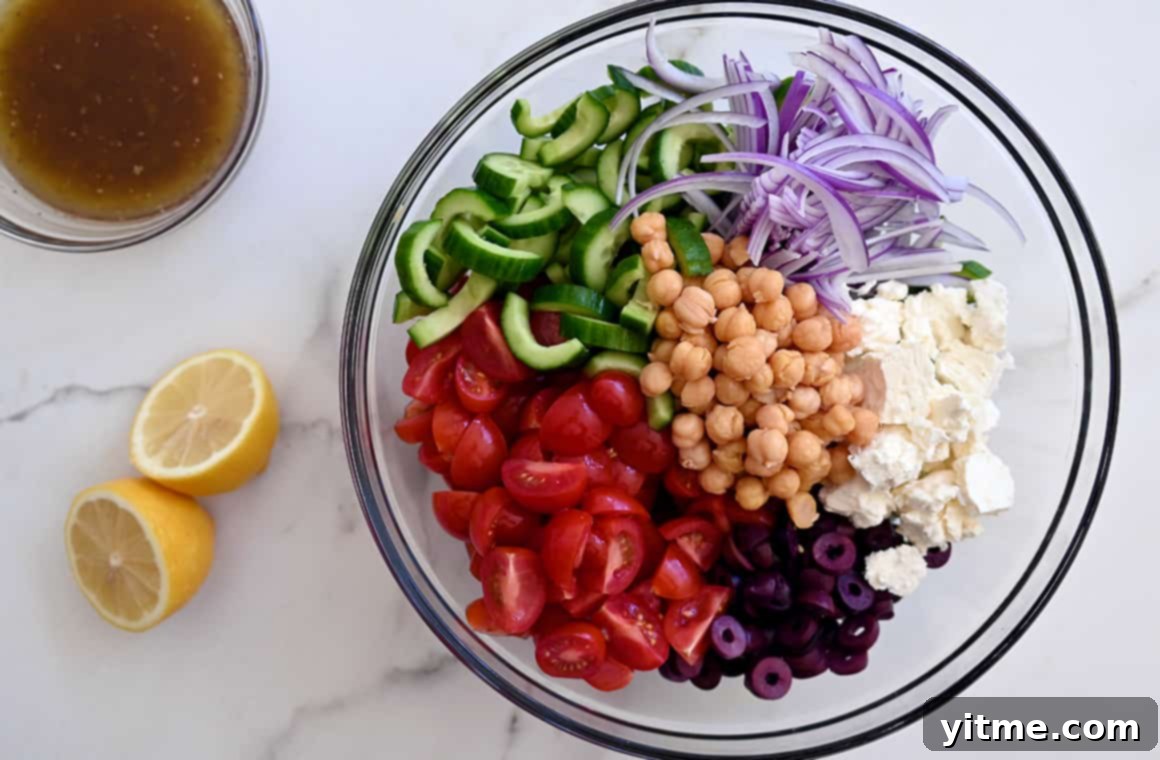 A clear bowl showcasing chickpeas, feta cheese, black olives, sliced red onions, halved cherry tomatoes, and sliced cucumbers