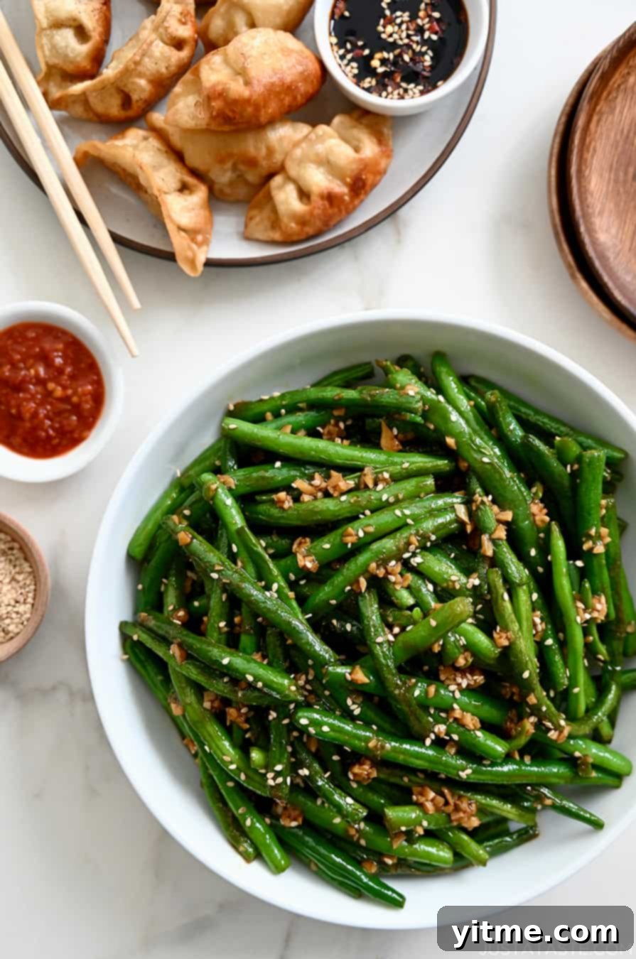 A white bowl containing Chinese Garlic Green Beans next to a plate with potstickers and chopsticks