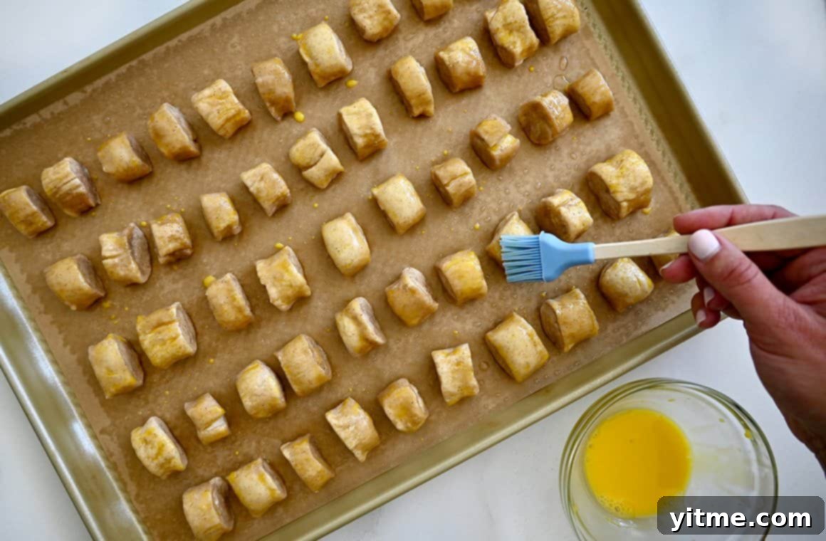 A top-down view of unbaked pretzel bites on a parchment paper-lined baking sheet being brushed with egg wash