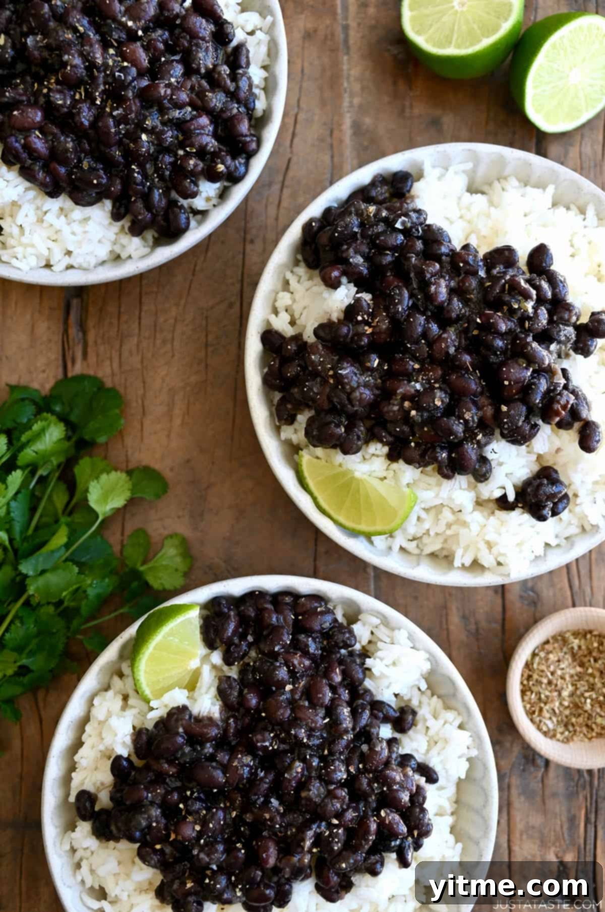 Three plates containing white rice topped with Cuban black beans and a lime wedge next to a bunch of cilantro.