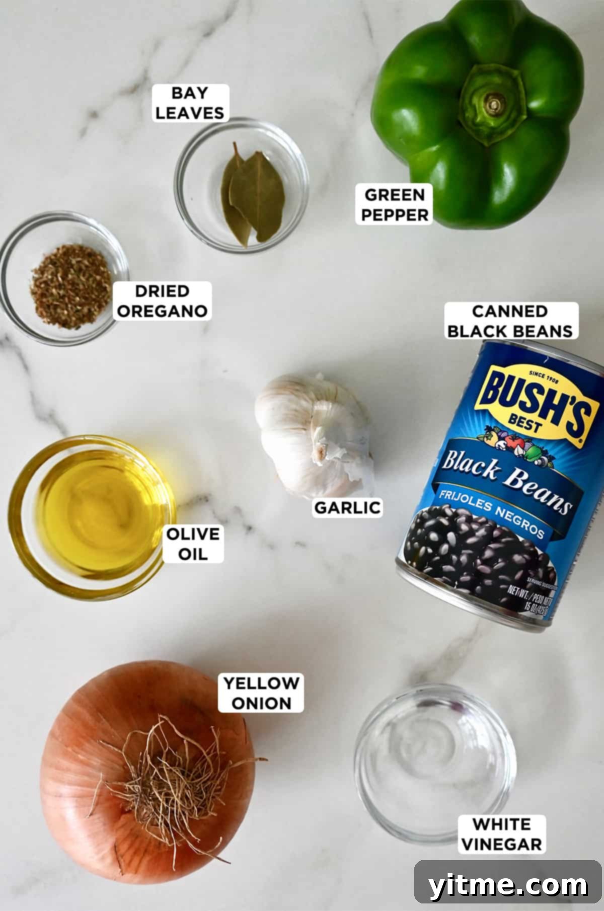A can of black beans next to a head of garlic, a yellow onion, and small bowls containing olive oil, dried oregano, bay leaves, green pepper and white vinegar.