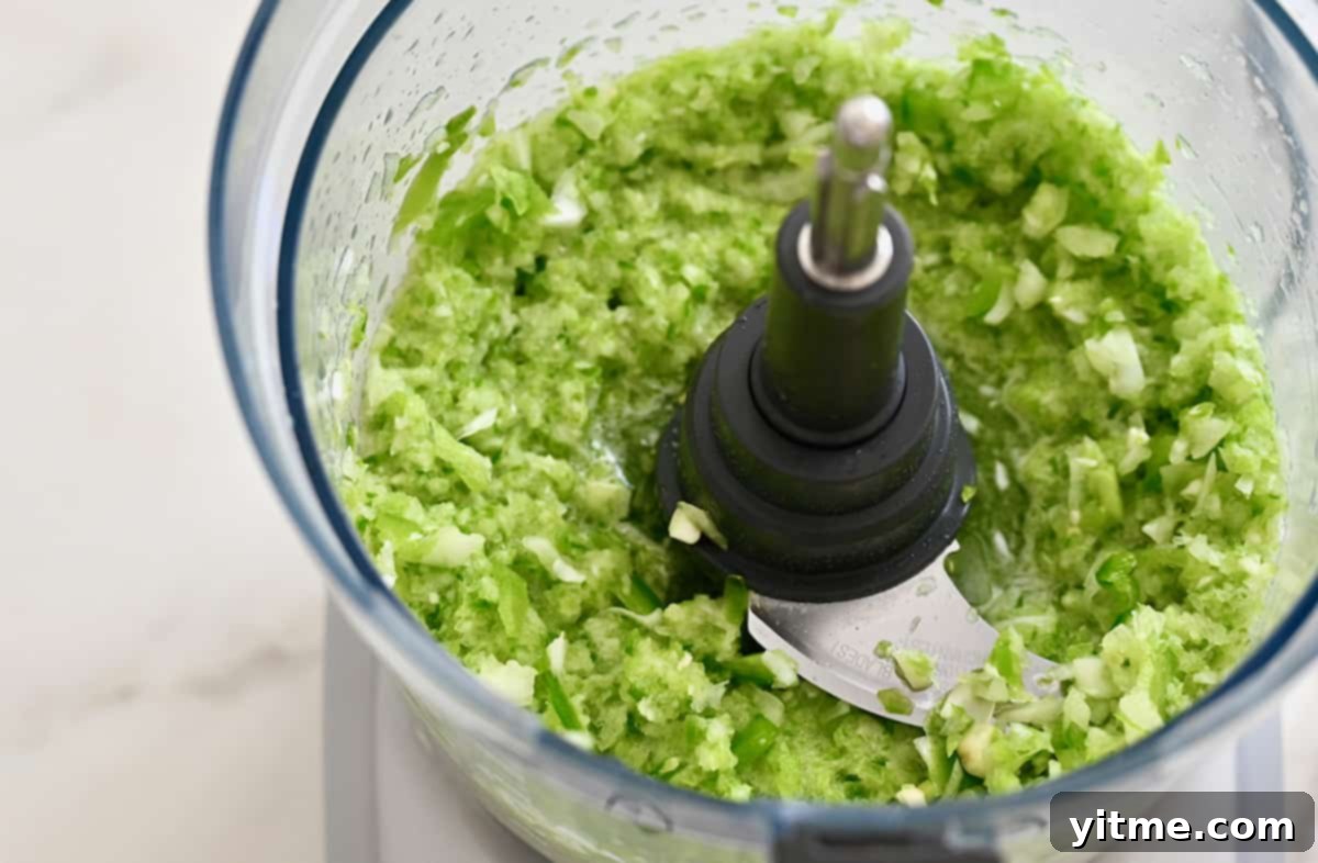 A food processor bowl containing sofrito, which is a mix of green bell pepper, onion and garlic.