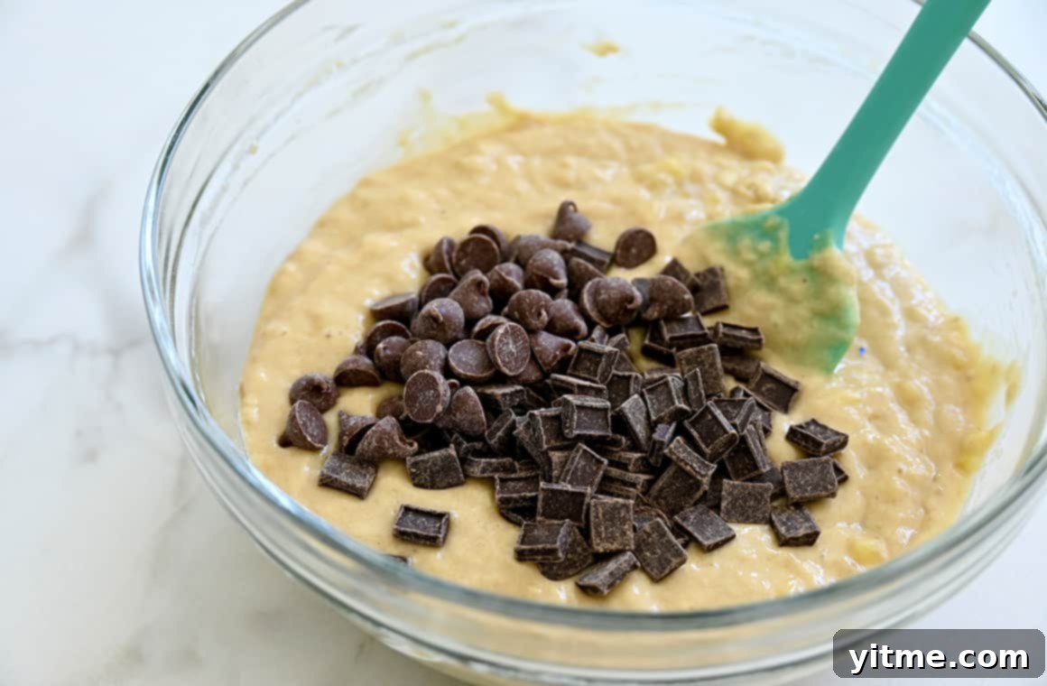 A teal spatula rests in a clear bowl containing batter, chocolate chips and chocolate chunks