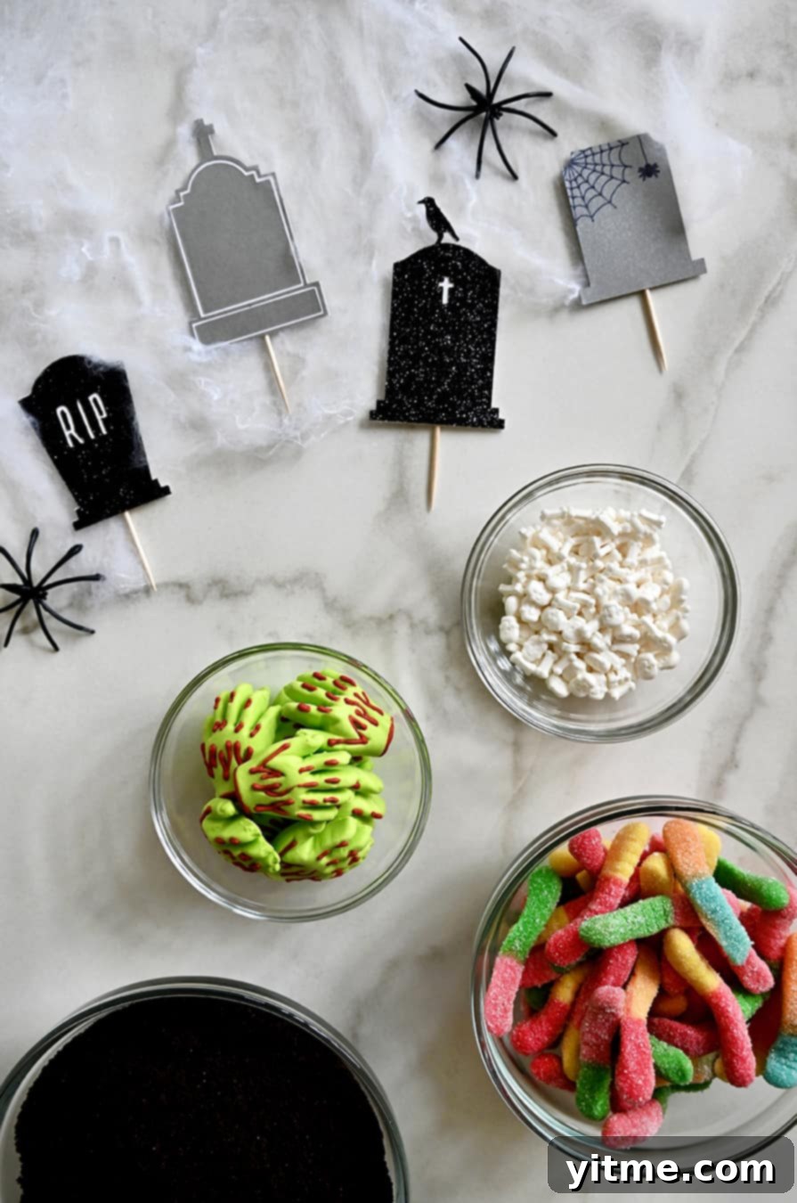 Overhead view of tombstone cake toppers alongside small bowls of candy bones, gummy worms, bloody-green gummy hands, and crushed Oreos for making Halloween Dirt Cups