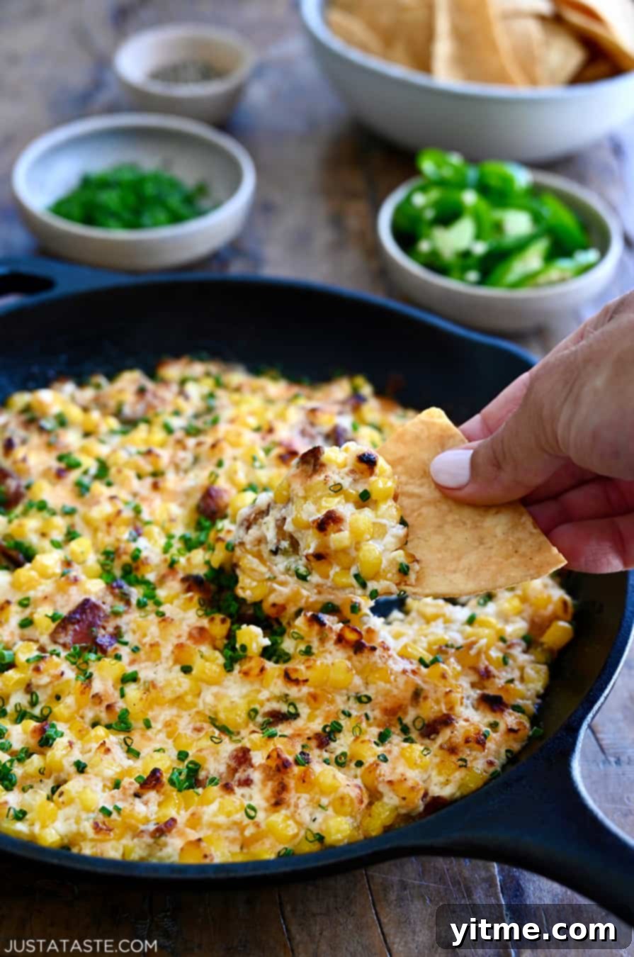 A hand dipping a crunchy tortilla chip into the warm, cheesy Jalapeño Corn Dip with Bacon in a cast iron skillet, emphasizing its delicious texture
