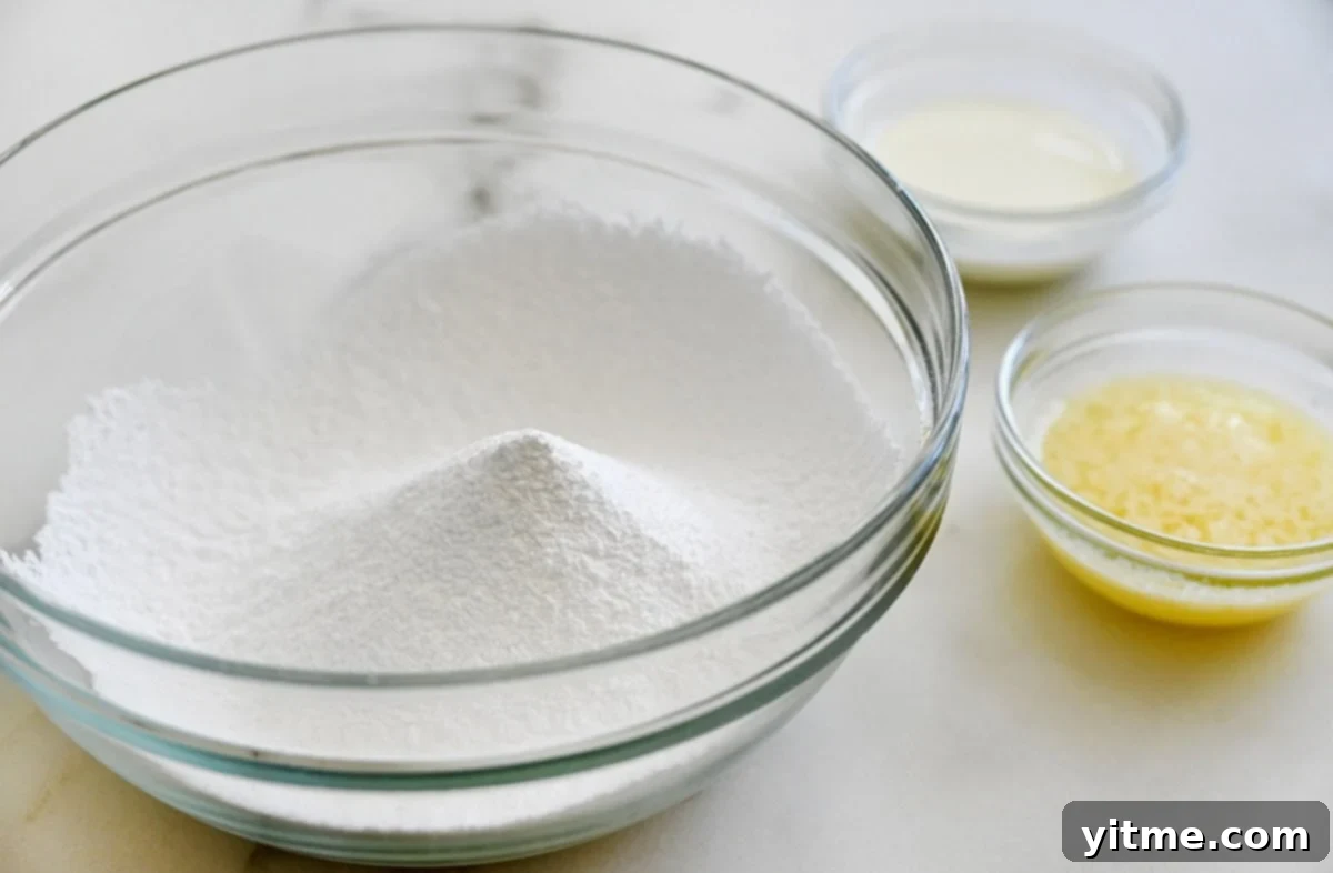 Silky Smooth Sugar Cookie Icing 4 A clear bowl containing confectioners’ sugar next to two small bowls containing melted butter and whole milk, ready for mixing.