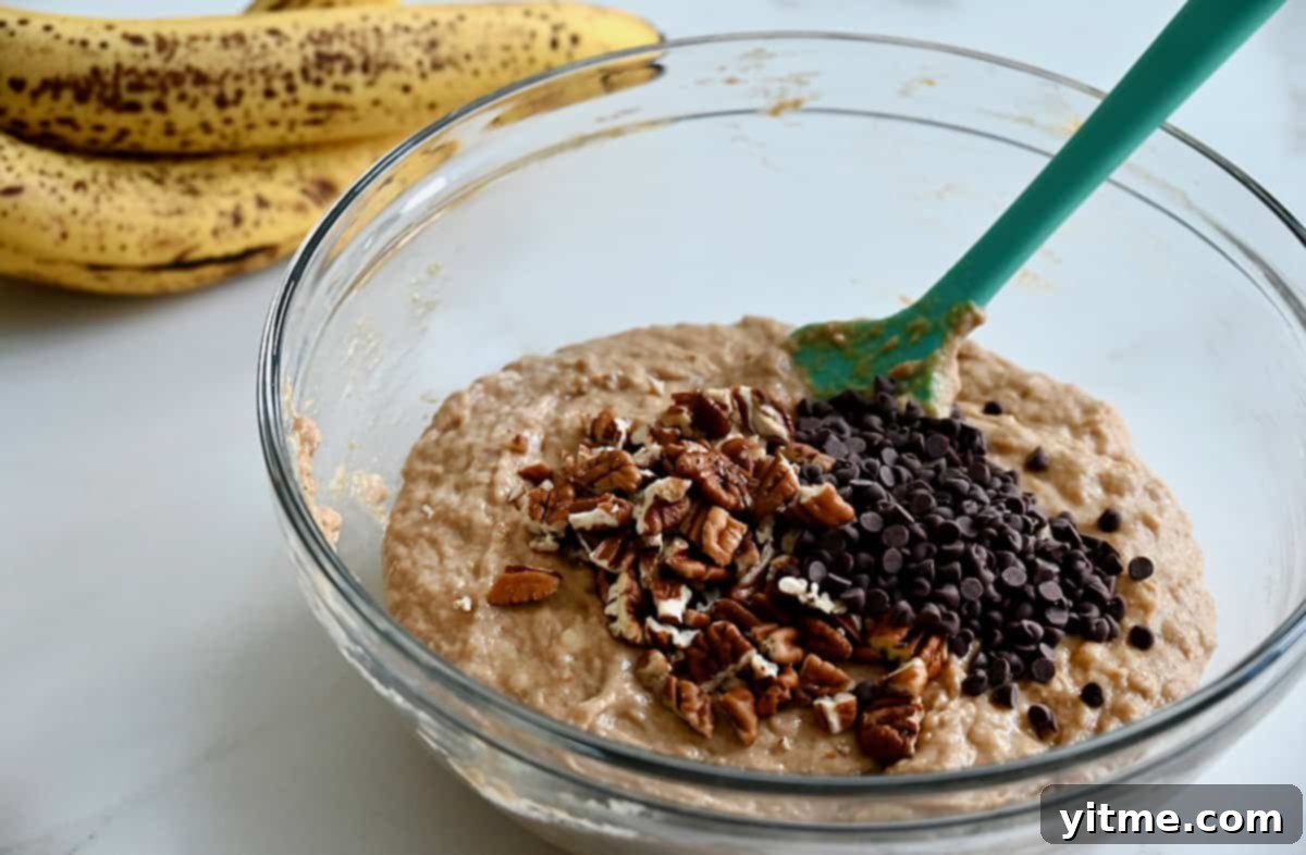 Whole wheat banana bread batter topped with mini chocolate chips and chopped pecans in a glass bowl next to a bunch of overripe bananas.