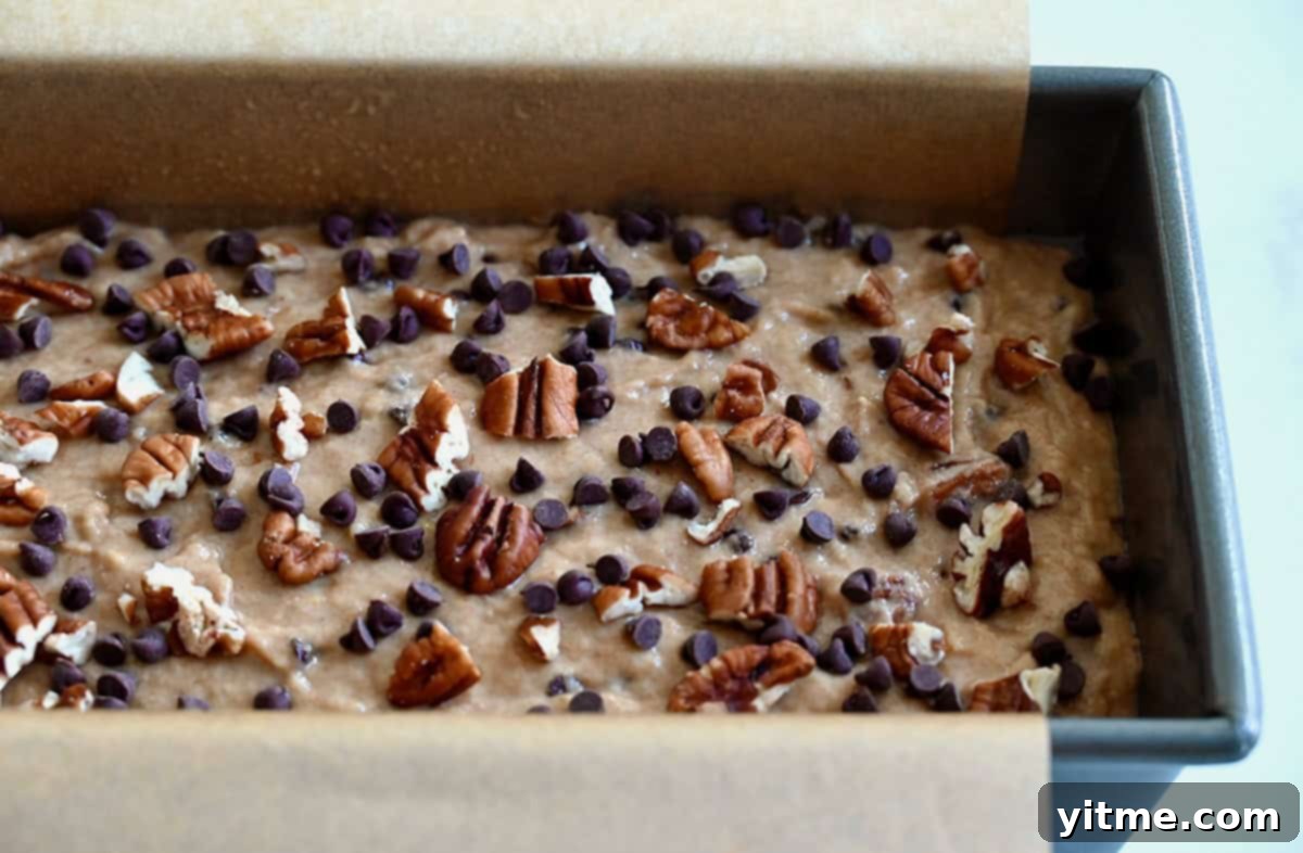 Whole wheat banana bread batter topped with mini chocolate chips and chopped pecans in a parchment paper-lined loaf pan.