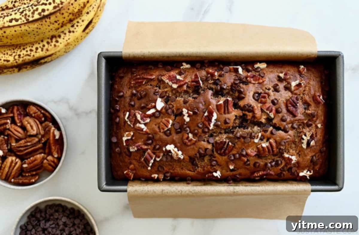 Healthy wheat banana bread in a loaf pan lined with parchment paper next to a bunch of overripe bananas and a small bowl containing mini chocolate chips.