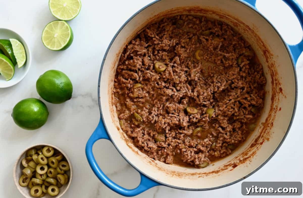 A large stockpot containing cooked ground beef next to limes and a small bowl containing sliced green olives.