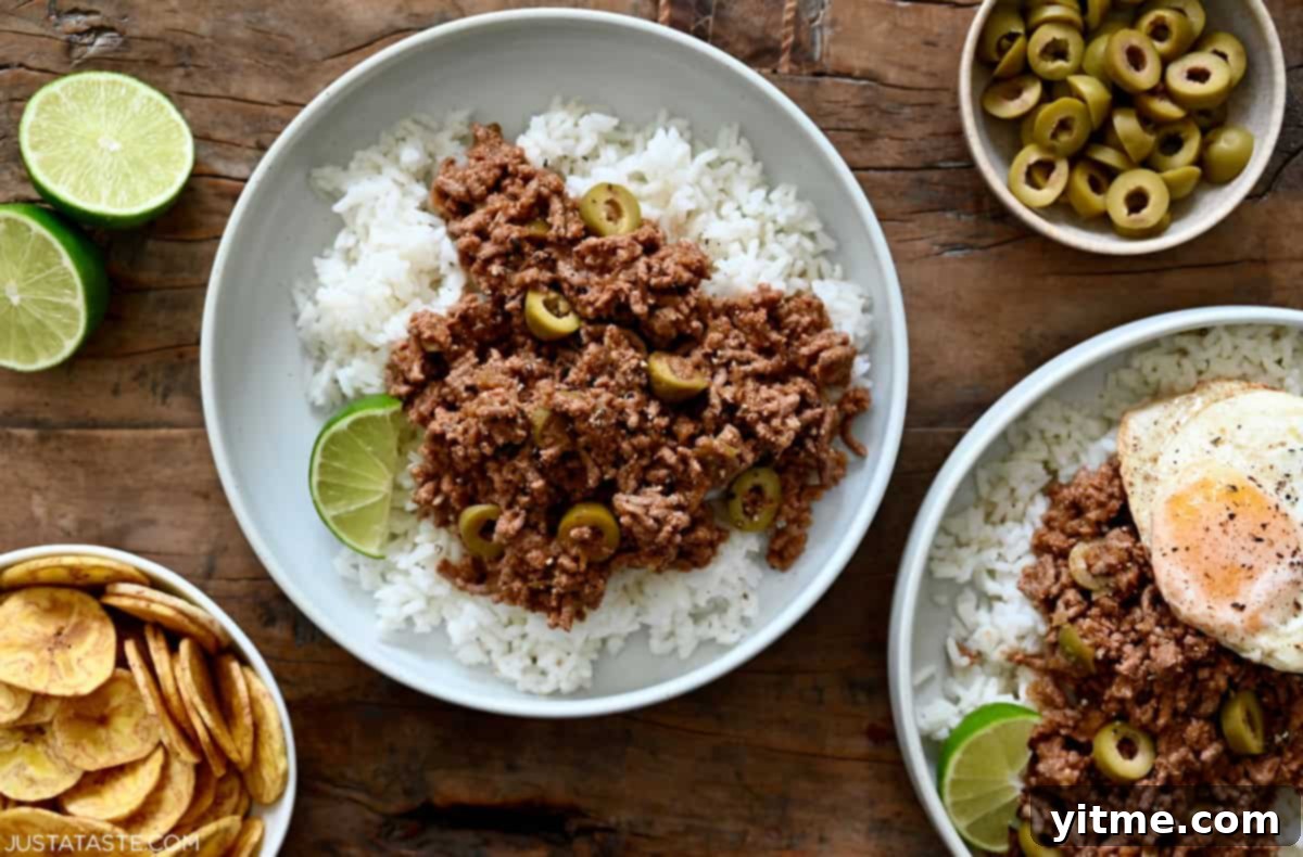Cuban picadillo (ground beef) with green olives and raisins atop steamed white rice in a bowl next to a small dish containing plantain chips.
