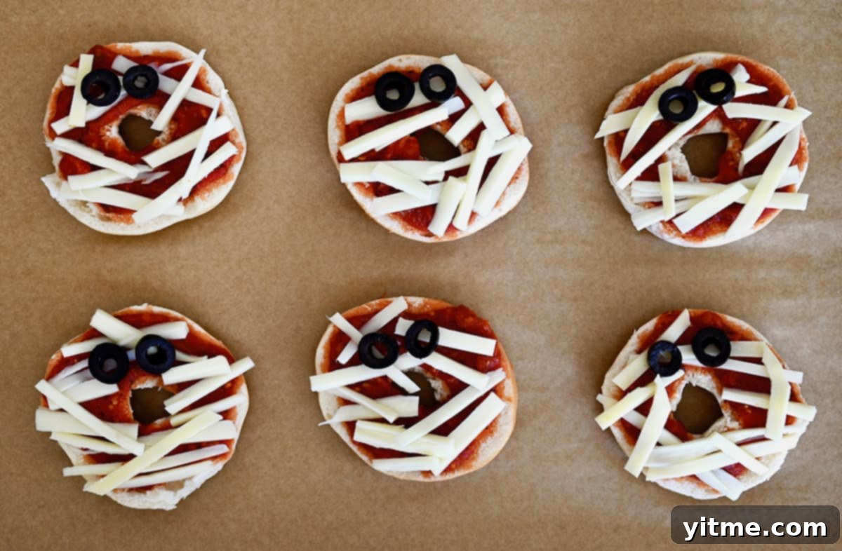 Six unbaked mummy pizza bagels on a parchment-lined baking sheet.