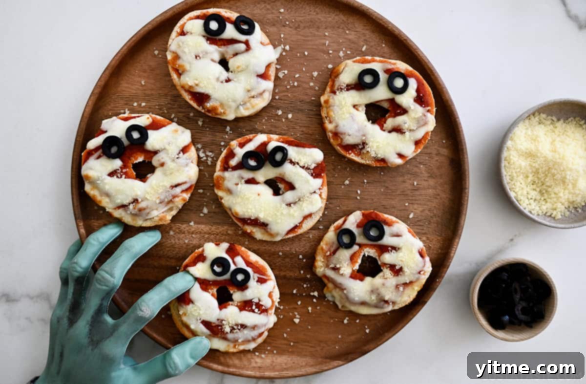 Six mummy pizza bagels on a wooden plate with small bowls of ingredients. A fake monster's hand is reaching over.