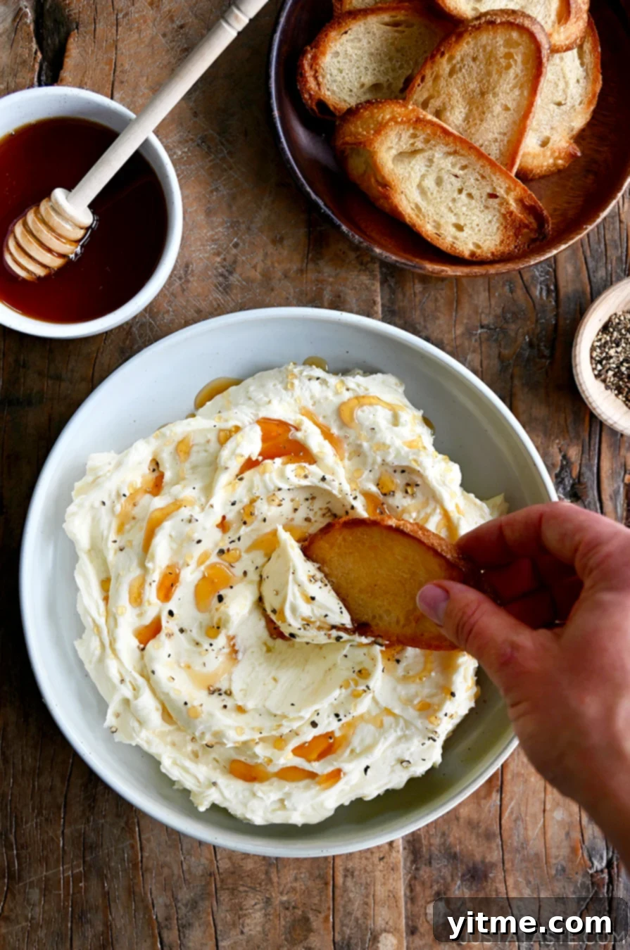 A hand dips a toasty baguette into whipped Brie that's been garnished with cracked black pepper and drizzled with honey.