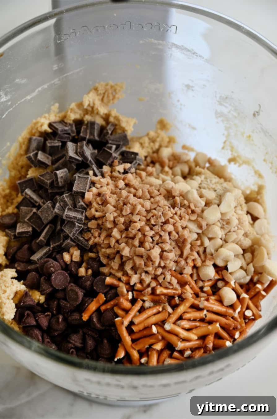 A clear bowl of a stand mixer filled with a vibrant assortment of cookie mix-ins: golden toffee bits, jagged crushed pretzel sticks, rich chocolate chunks, perfectly chopped macadamia nuts, and classic semisweet chocolate chips, ready to be folded into dough.