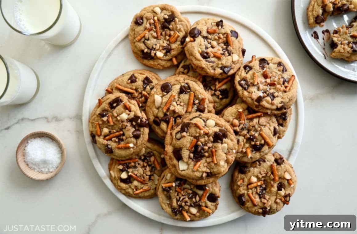 A delightful pile of Kitchen Sink Cookies, generously loaded with chocolate, chopped nuts, buttery toffee bits, and salty pretzels, perfectly arranged on a plate alongside two tall glasses of fresh milk, inviting a delicious treat.