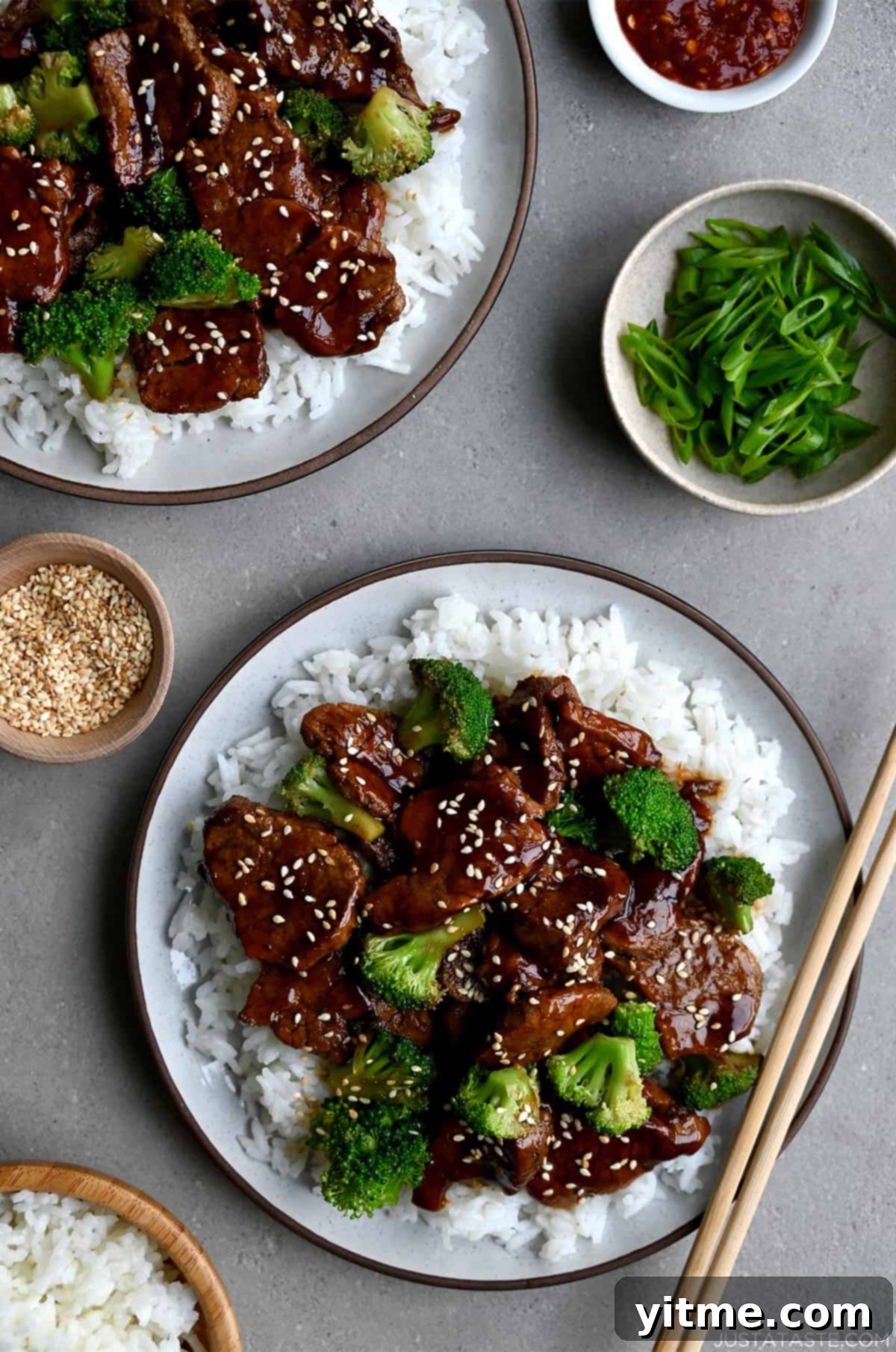 Quick Caramelized Pork and Broccoli over a bed of rice on a plate with chopsticks next to a small bowl containing sesame seeds and a bowl containing broccoli florets.