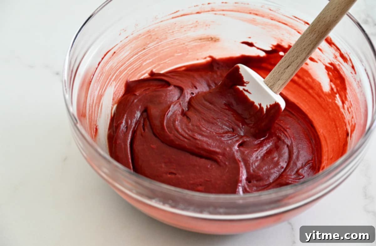 Ruby Velvet Brownie Cookies 5 Vibrant red velvet brownie batter in a large clear glass bowl, being mixed with a spatula, showcasing its rich color and smooth consistency, ready to be combined with the cookie dough.