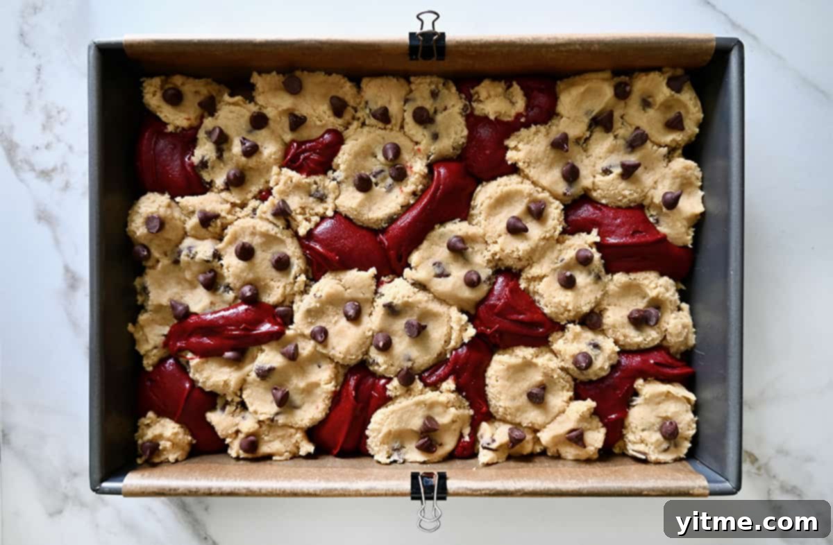 Ruby Velvet Brownie Cookies 6 A top-down view of beautifully marbled unbaked red velvet brownie and chocolate chip cookie dough swirled together in a parchment paper-lined baking pan, patiently waiting to be baked in the oven.